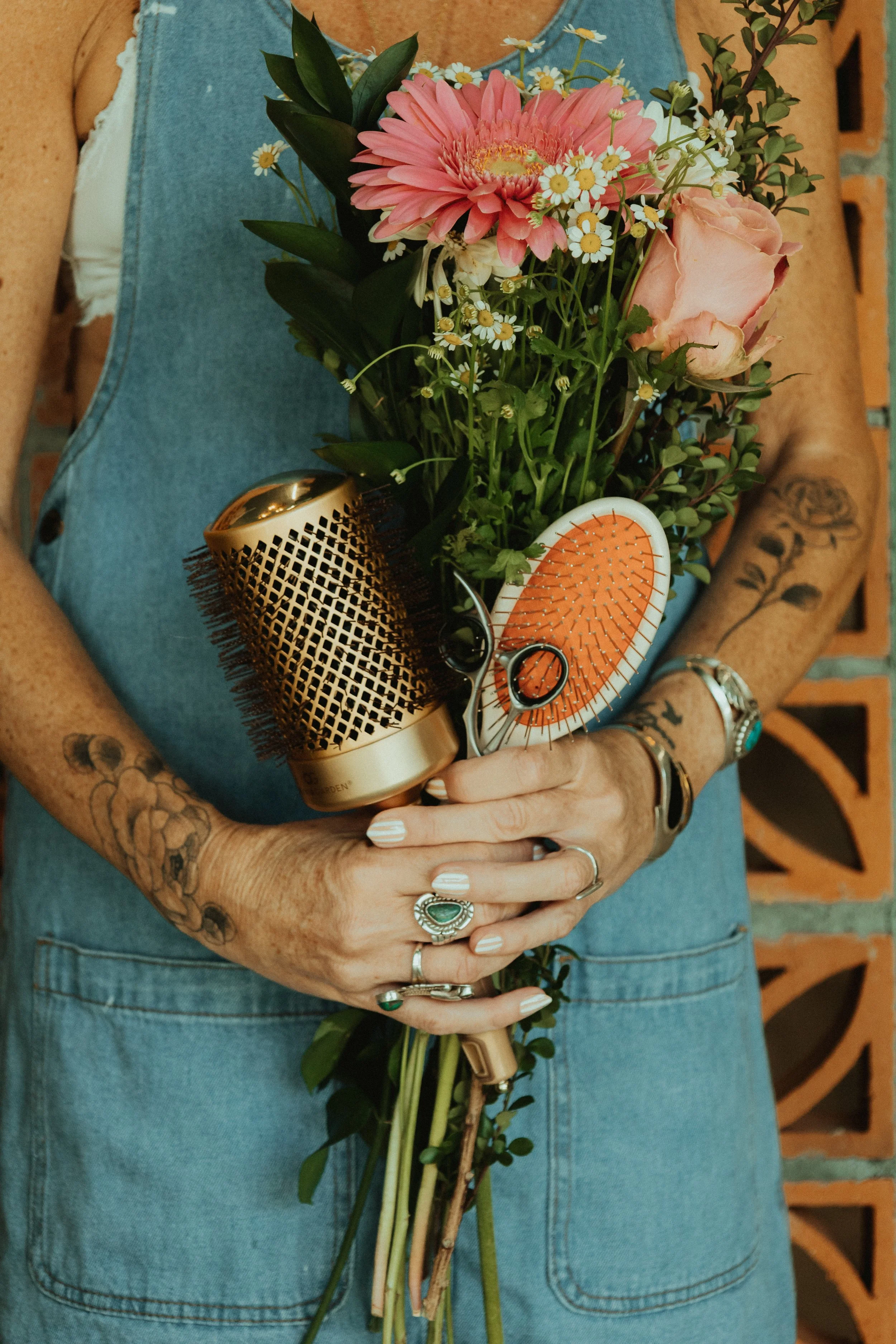 Person holding a bouquet of pink and white flowers, hairstyling tools, and jewelry, wearing a sleeveless denim dress and tattoos, with a brick wall background.