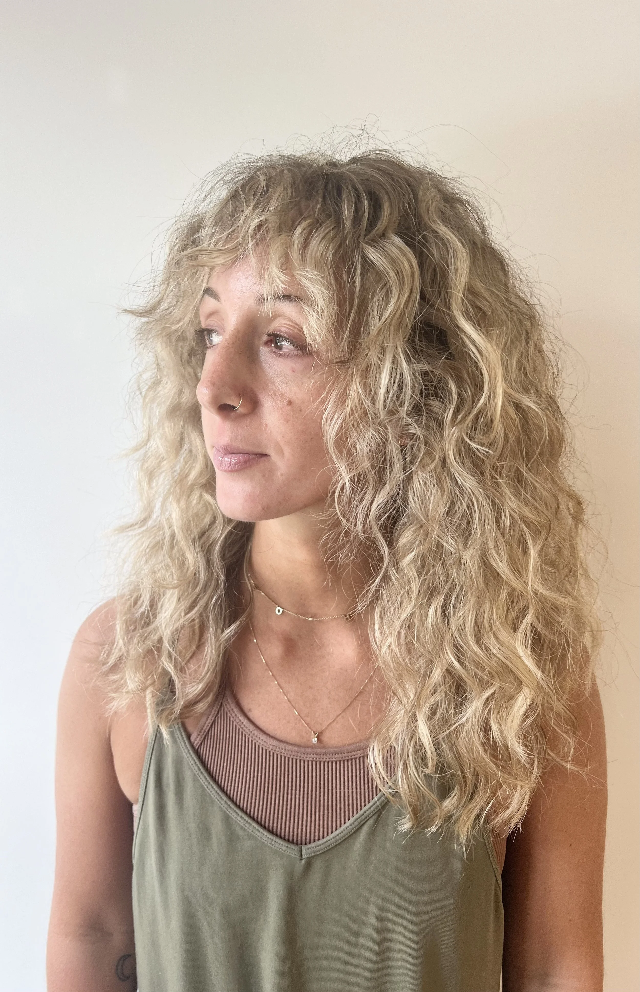 A woman with curly blonde hair wearing a green top with a layered necklace, looking to the side.