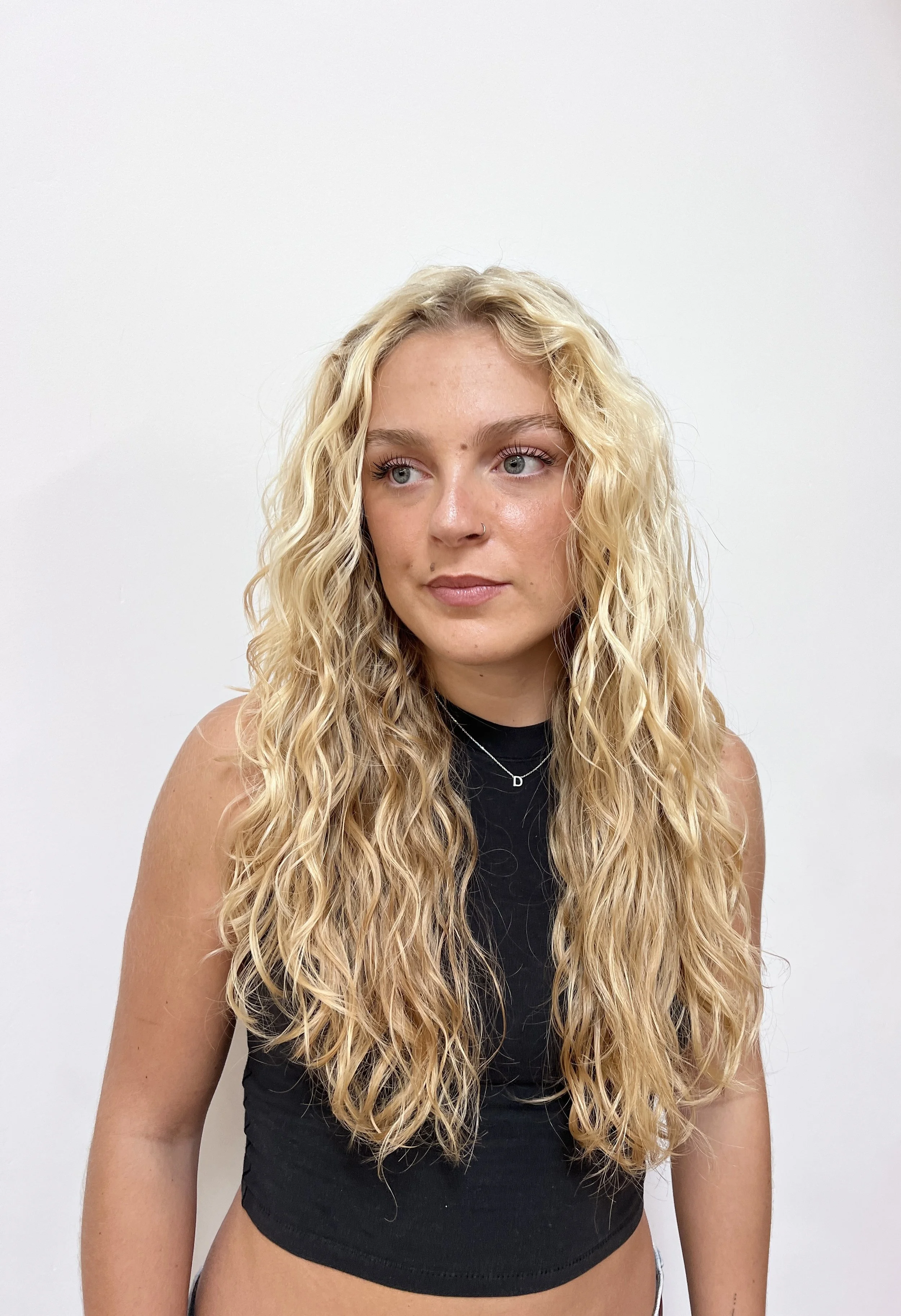 A young woman with long, curly blonde hair and blue eyes, wearing a black sleeveless top and a delicate necklace, standing against a plain white background.