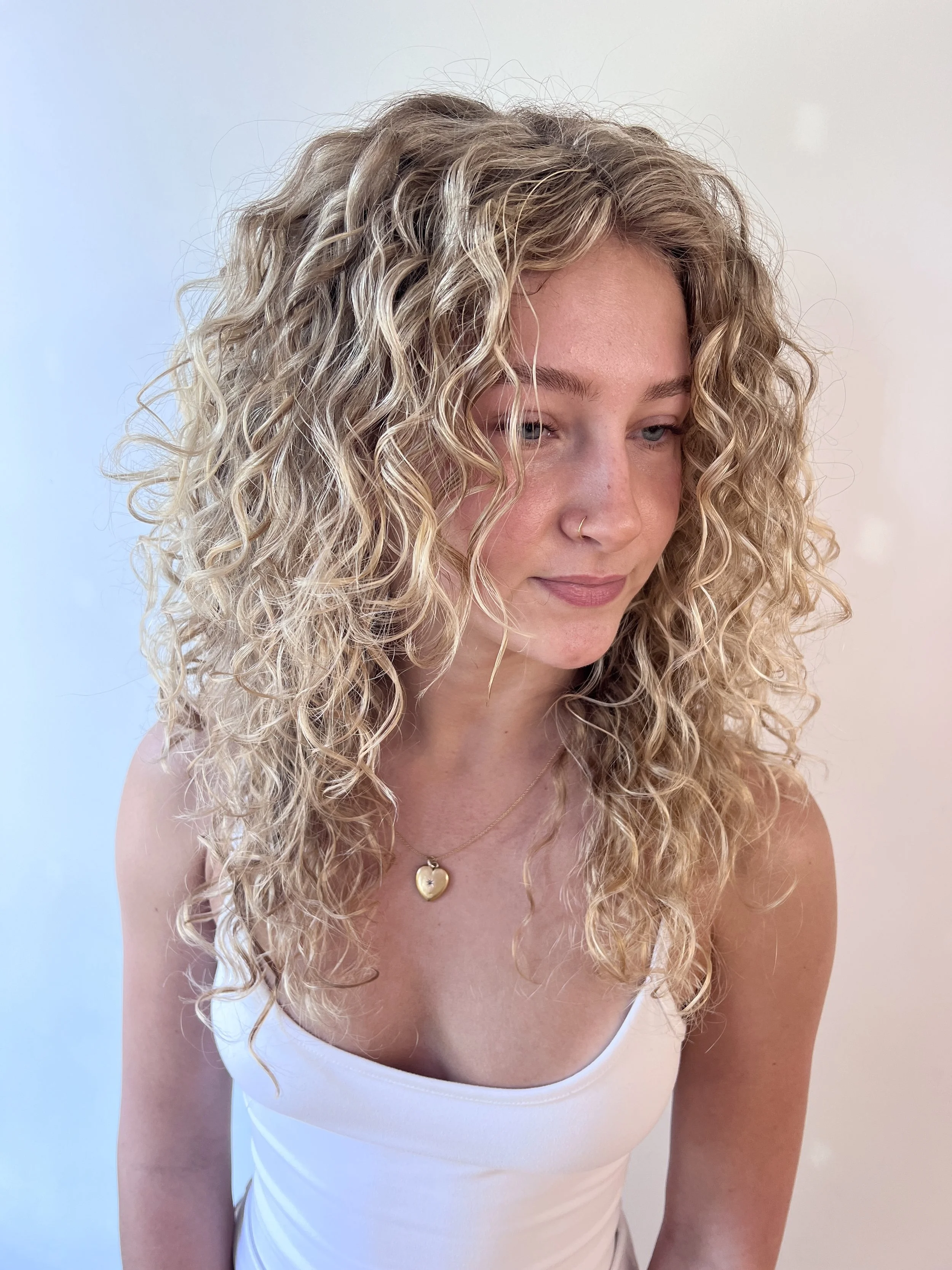 A young woman with curly blonde hair, wearing a white tank top, a gold heart-shaped necklace, and a nose ring, standing against a plain light-colored background.