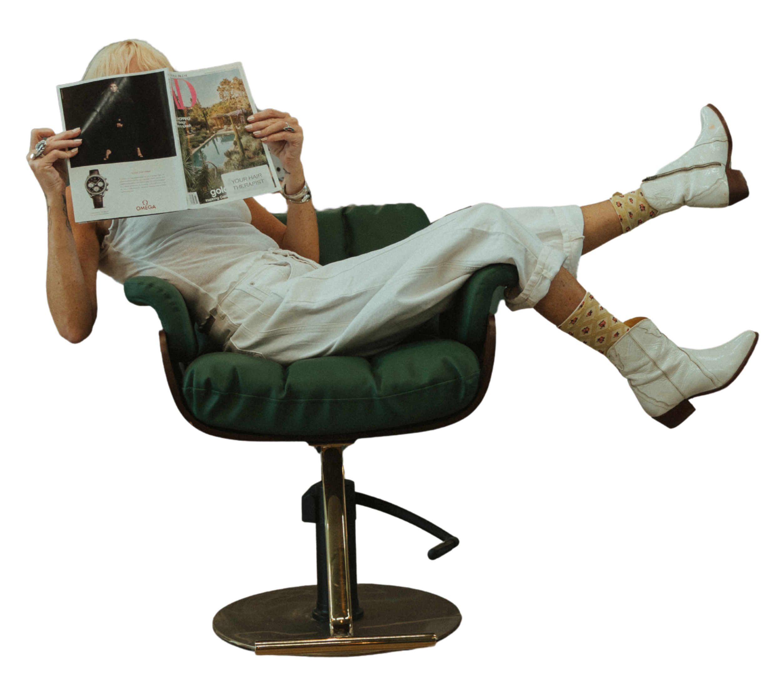A person with blond hair and white clothing lounging in a green armchair, reading a magazine with their legs extended, wearing white boots, beige socks with a pattern, and gold jewelry.