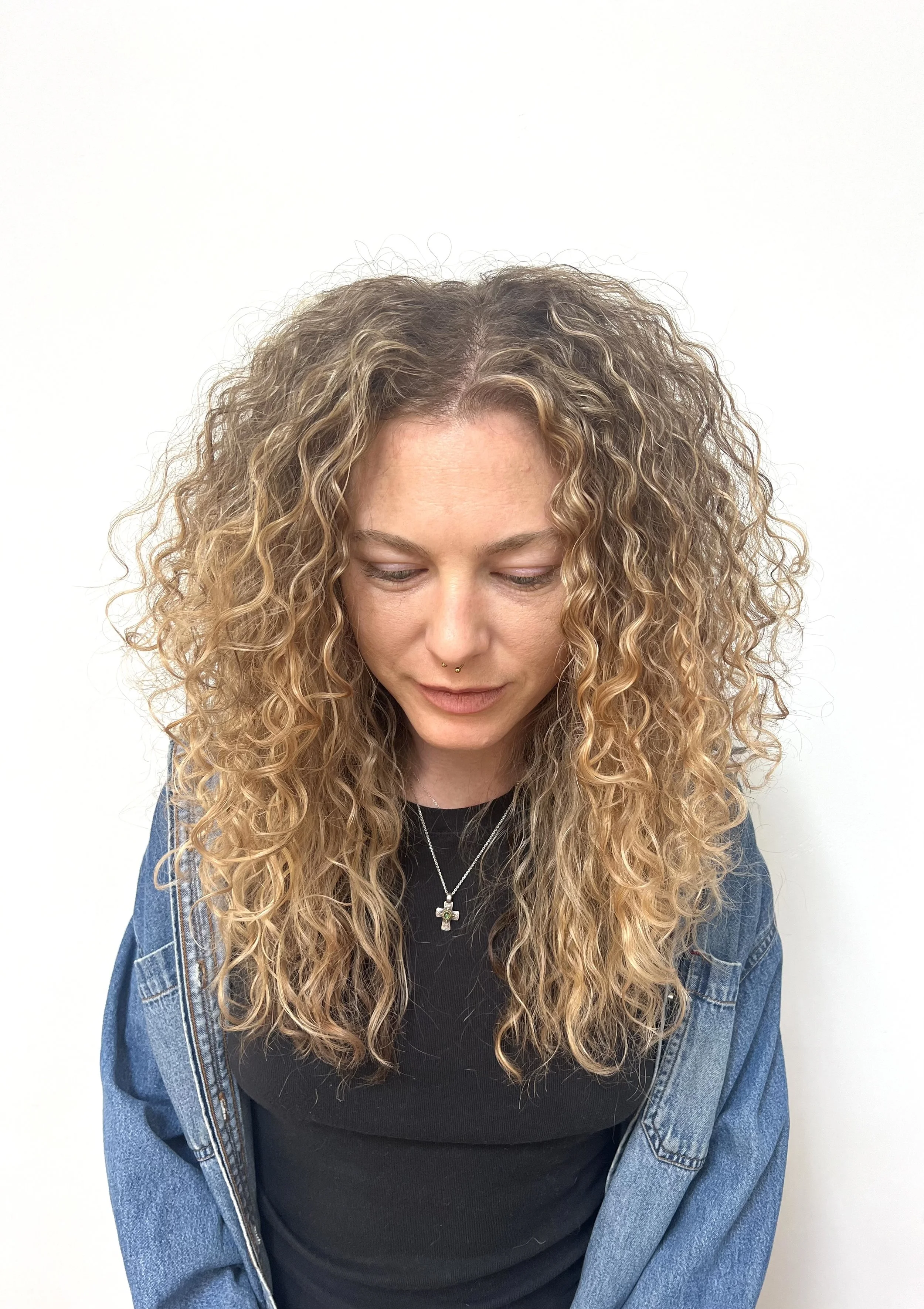 A woman with long, curly blond hair looking down, wearing a black shirt, denim jacket, and silver jewelry against a plain white background.