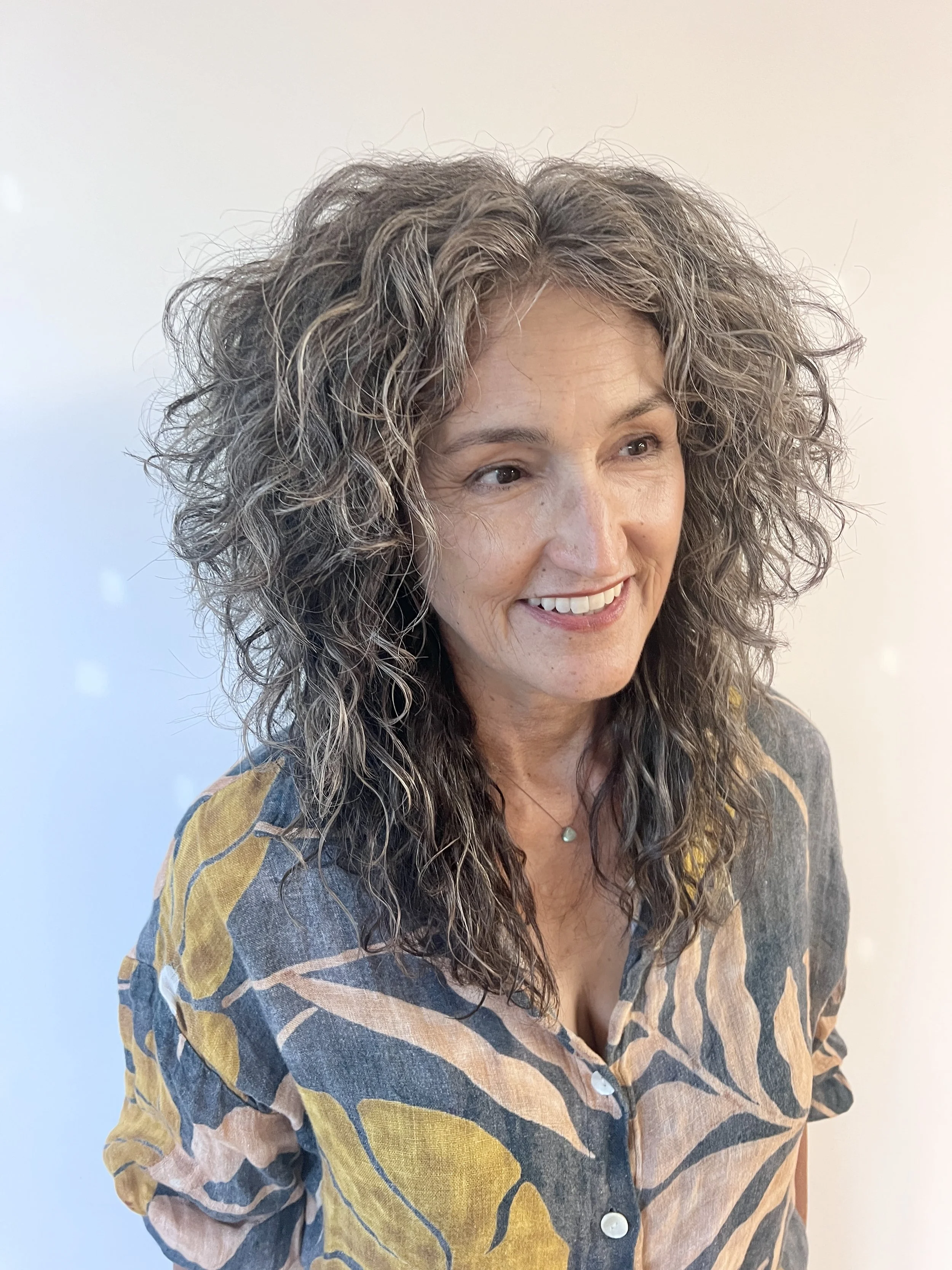 A woman with curly gray hair smiling, wearing a patterned shirt with yellow and blue leaves, standing against a light background.