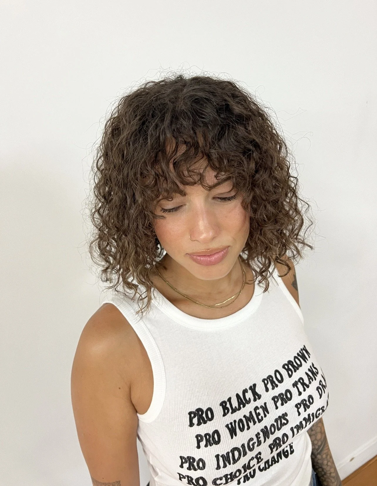 Woman with curly brown hair wearing a white tank top with black text, standing against a plain white background.