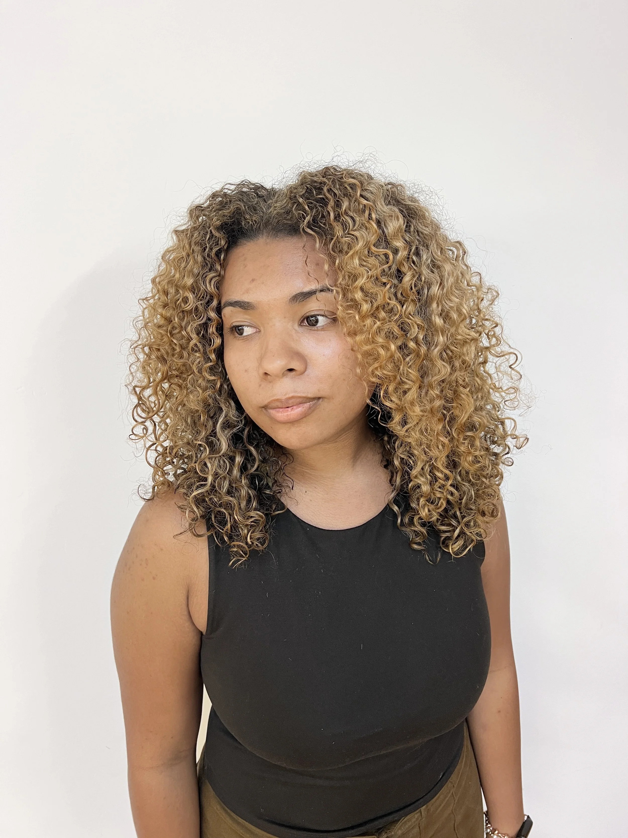 A woman with curly, blonde and brown hair wearing a black sleeveless top standing against a plain white wall.