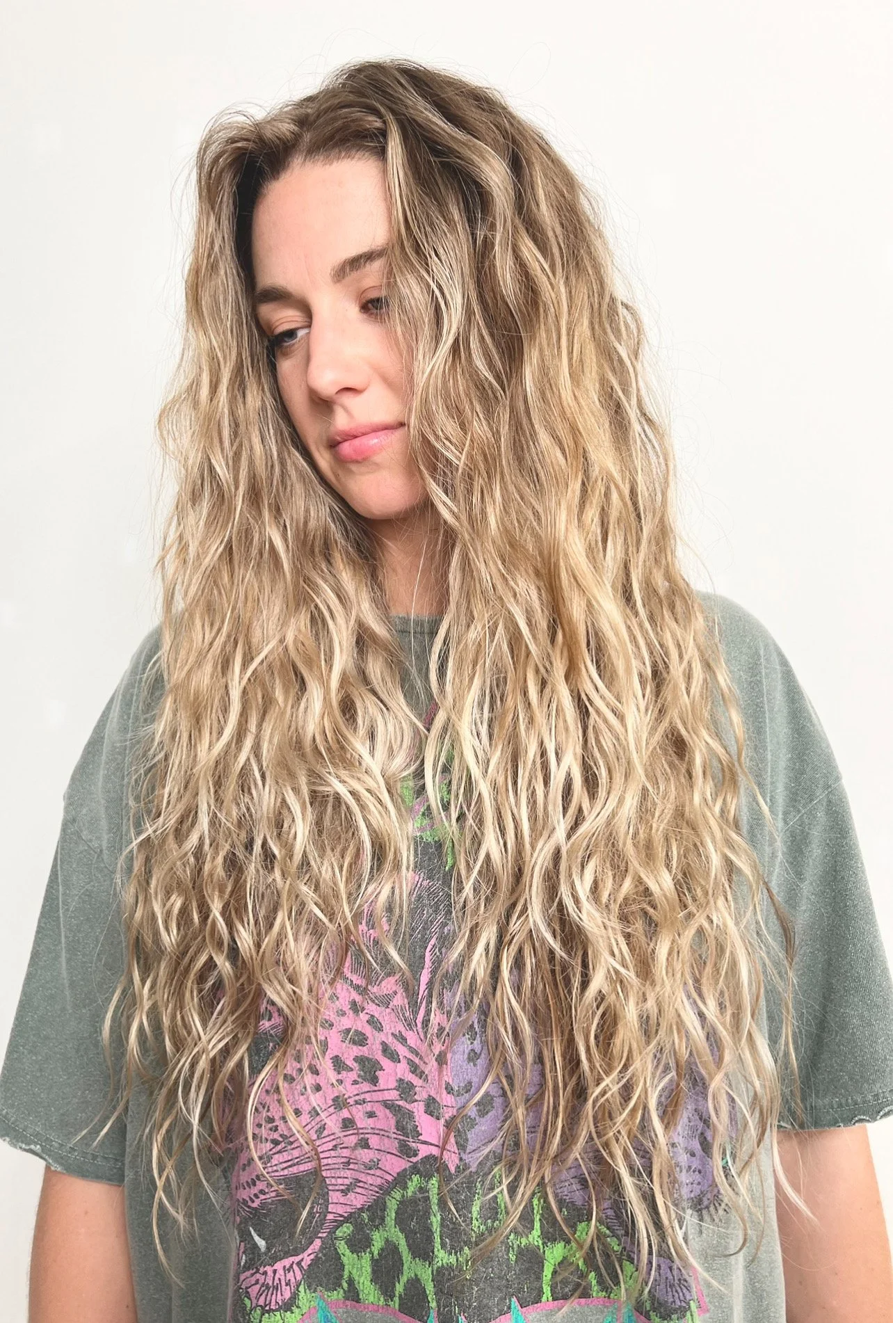 A woman with long, curly blonde hair wearing a graphic t-shirt against a plain white background.