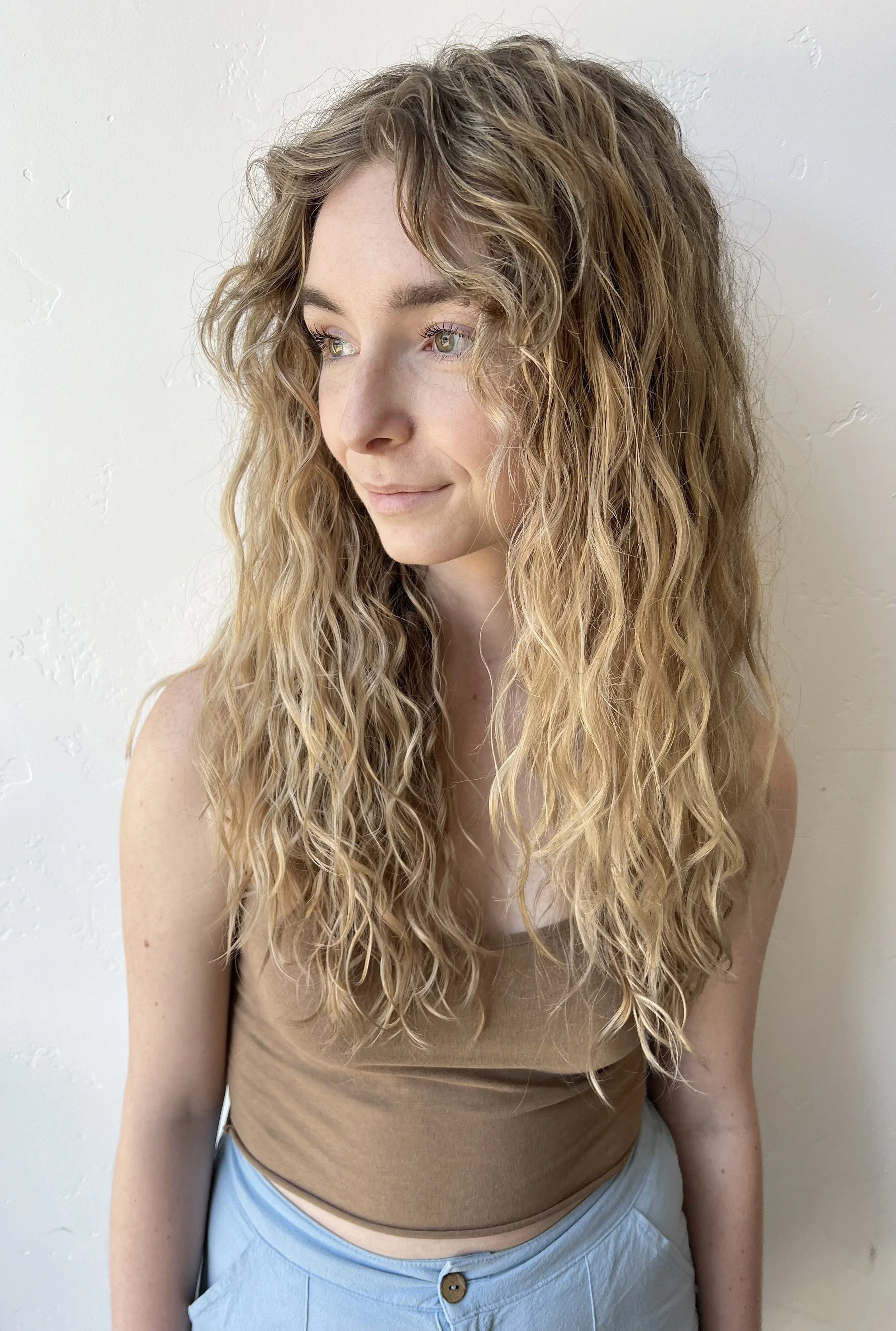 A young woman with curly blonde hair and light eyes looks to her left, standing against a light-colored wall.