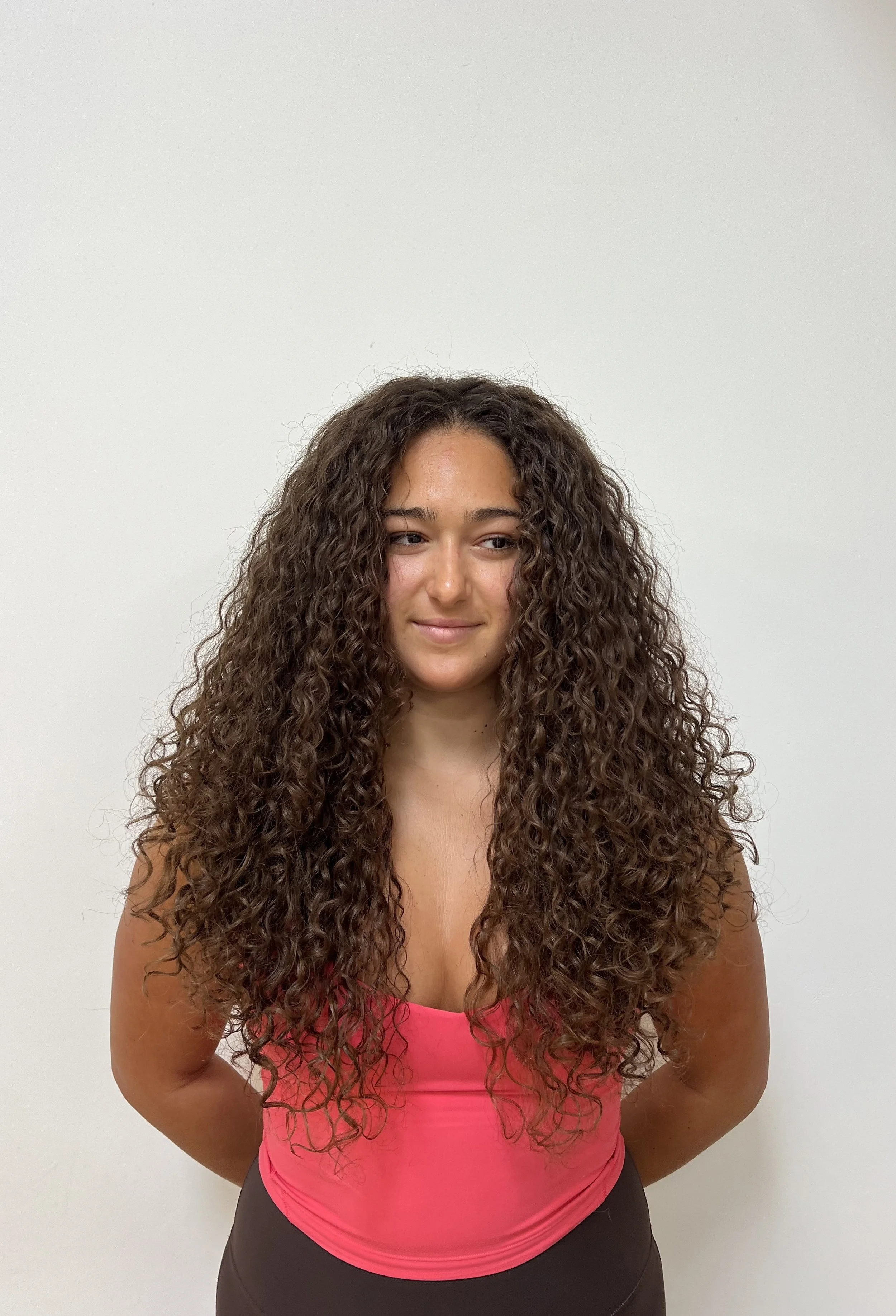Young woman with long curly brown hair wearing a pink top and black pants standing against a plain white wall.