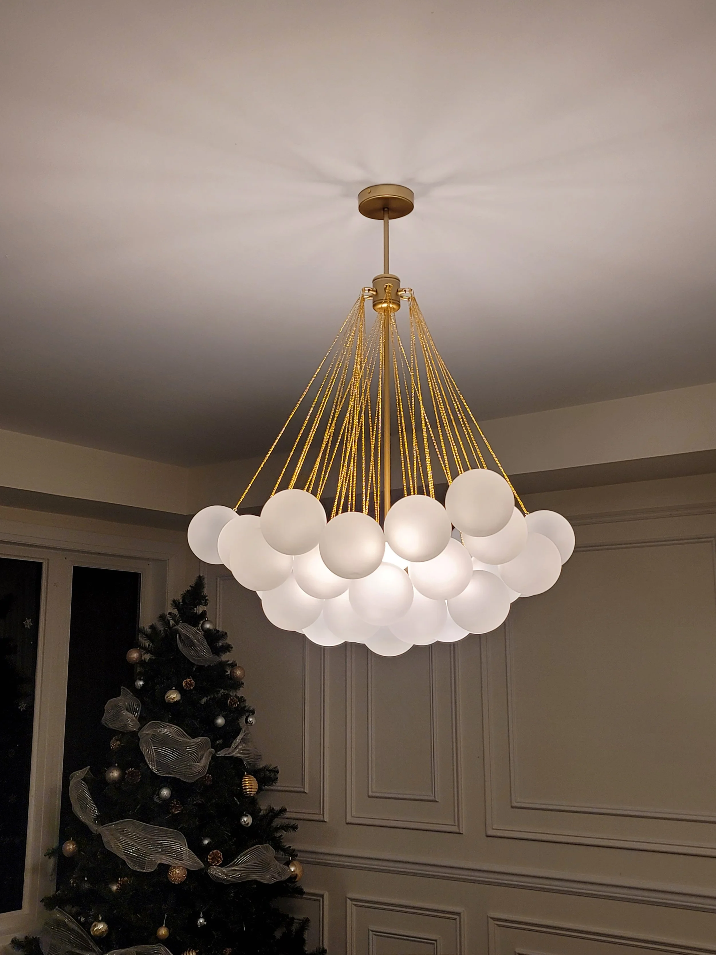 A modern chandelier with multiple white globe lights hanging from gold cords in a living room with a decorated Christmas tree.
