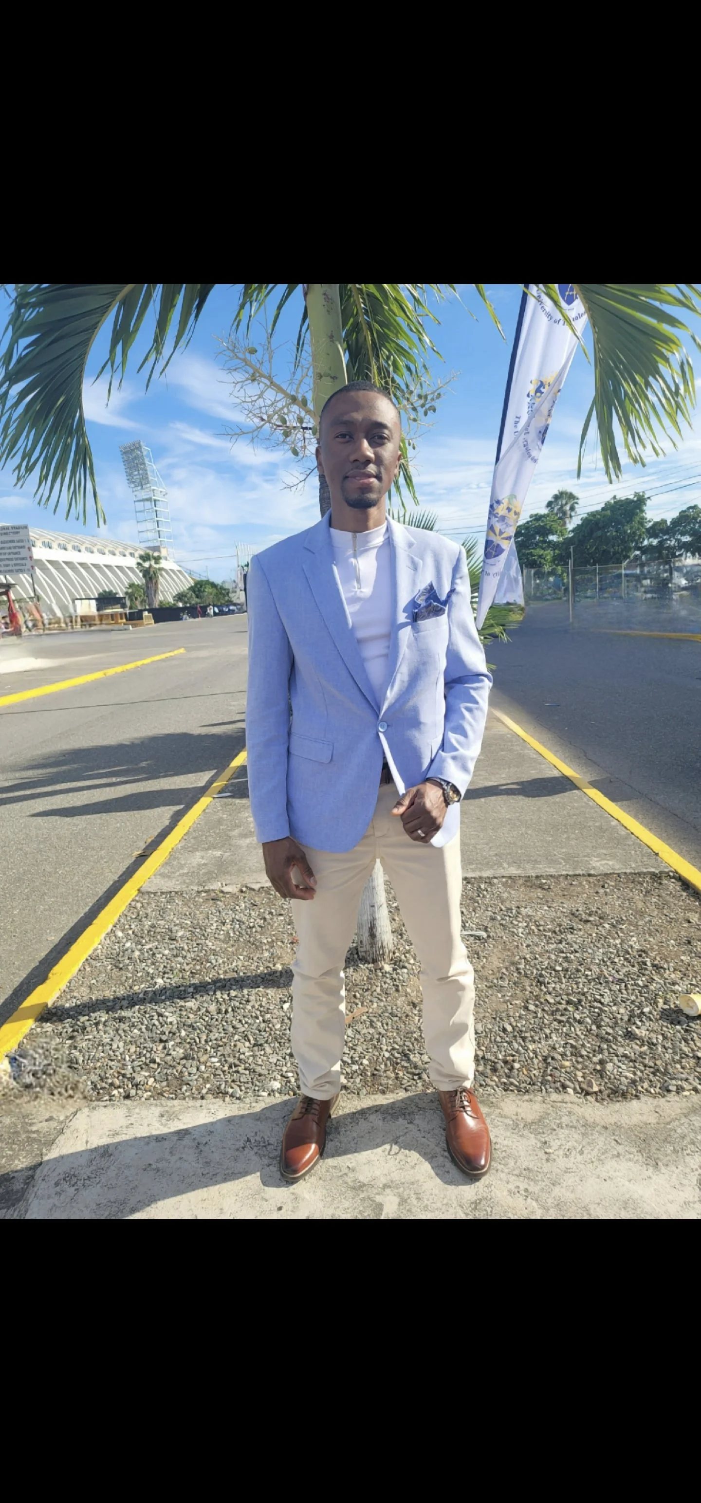 Man dressed in a light blue blazer, white shirt, beige pants, and brown shoes standing outdoors near a tree with palm leaves. Background includes a sporting stadium, flag, and a blue sky with some clouds.