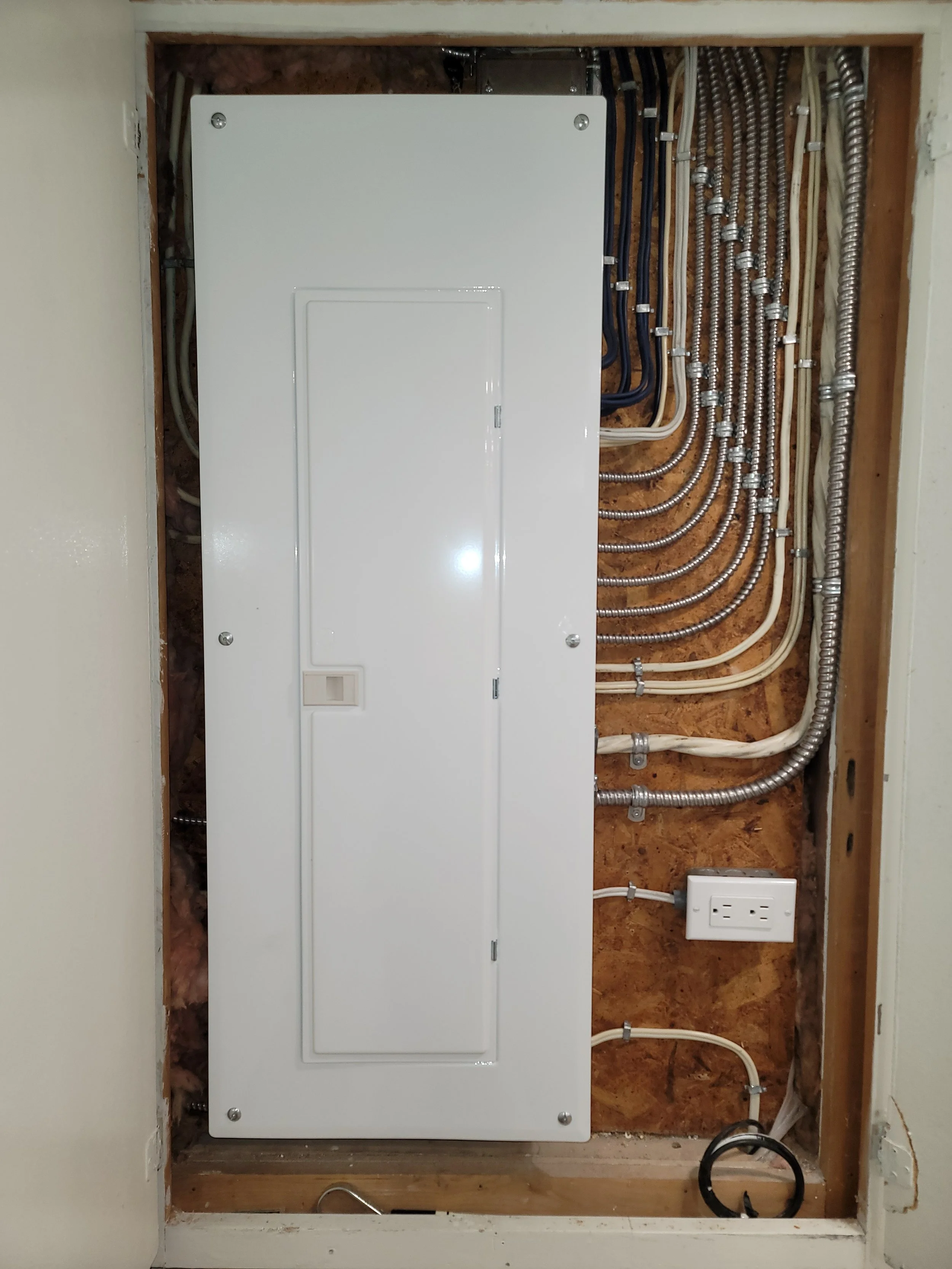 Electrical panel with wiring inside a wall cavity, including conduit and an electrical outlet.