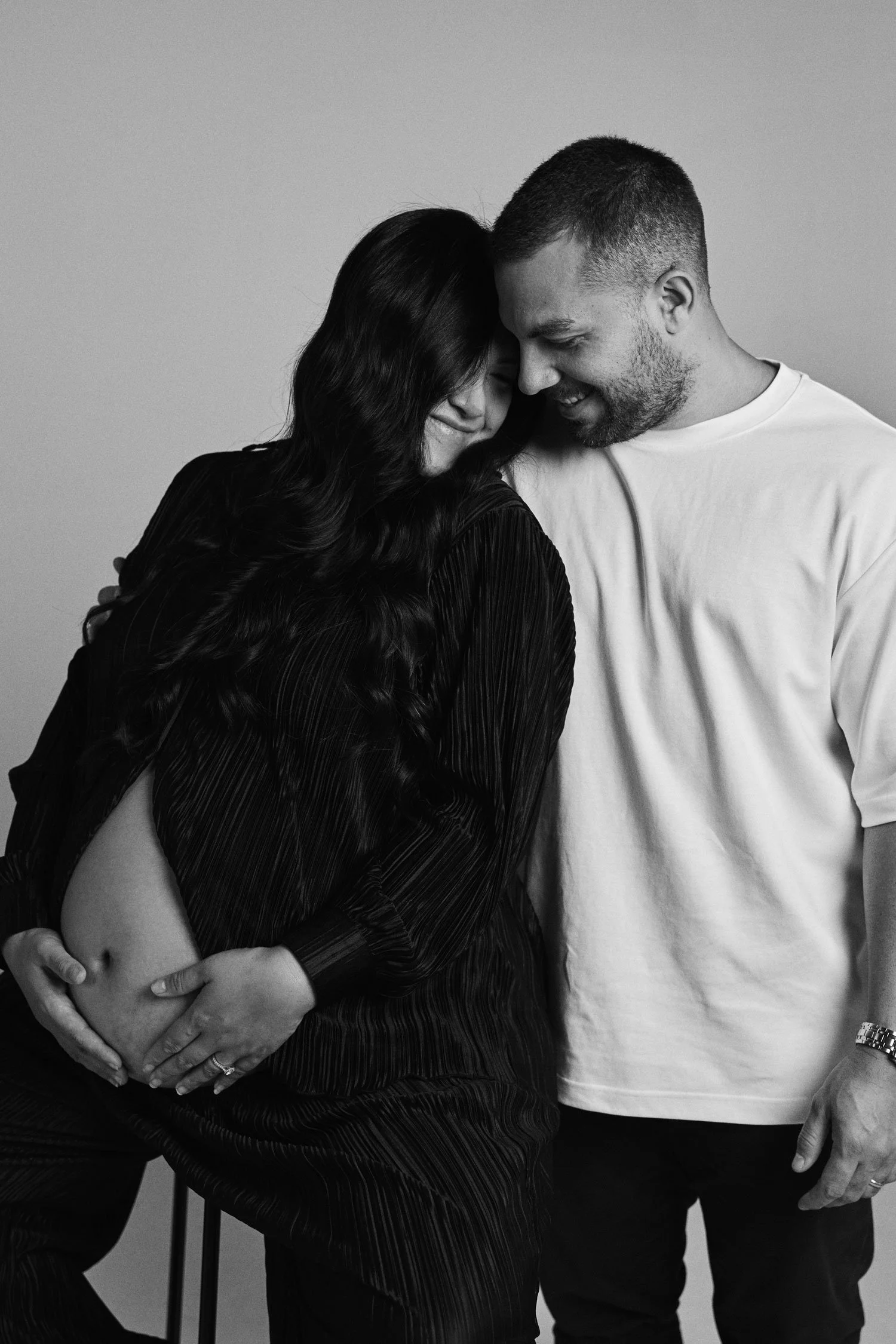 A pregnant woman with long, dark hair and a man with short hair and a beard, both smiling and touching foreheads, in a black and white photo.