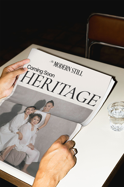 Person flipping through a fashion magazine titled 'The Modern Still Heritage' on a white table with a glass of water nearby.