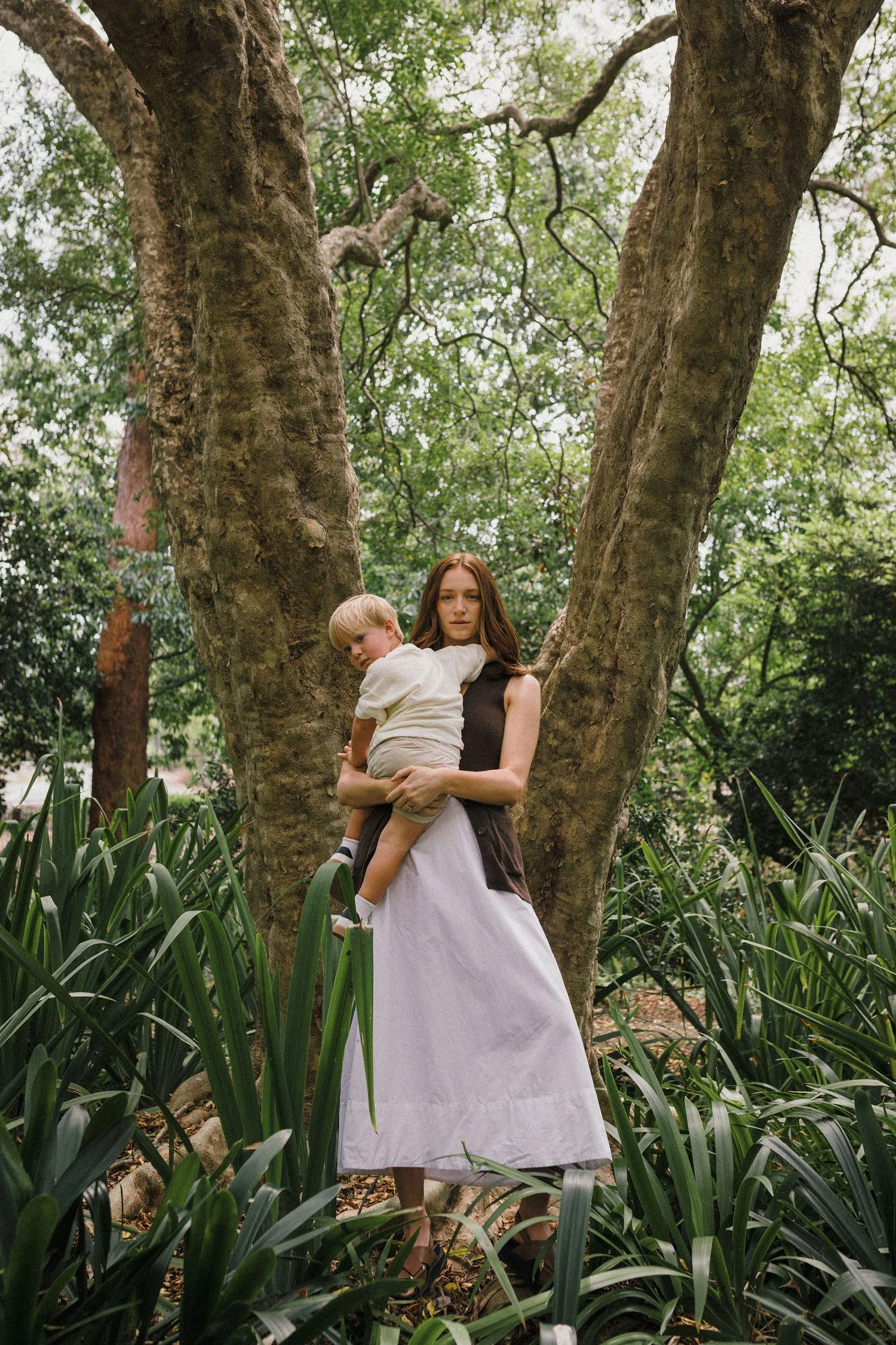 A woman with long reddish-brown hair wearing a sleeveless black top and white wide-leg skirt stands outdoors near a large tree, holding a young blonde boy wearing a white shirt and beige shorts in her arms. The scene is set in a lush, green wooded area with tall grass and dense foliage.