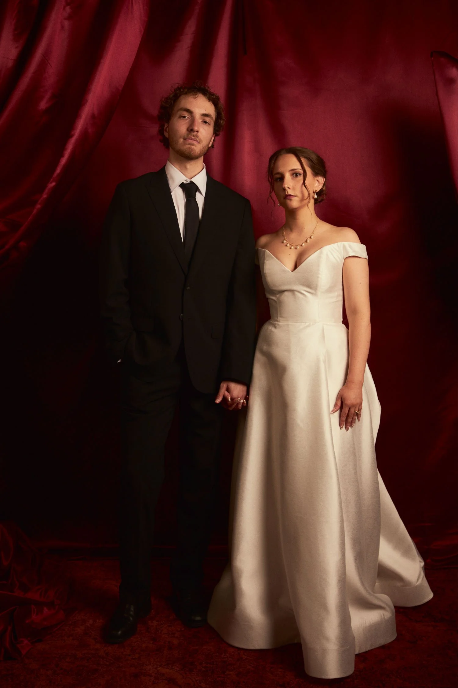 A man in a black tuxedo and a woman in a white wedding dress holding hands and standing against a red curtain background.