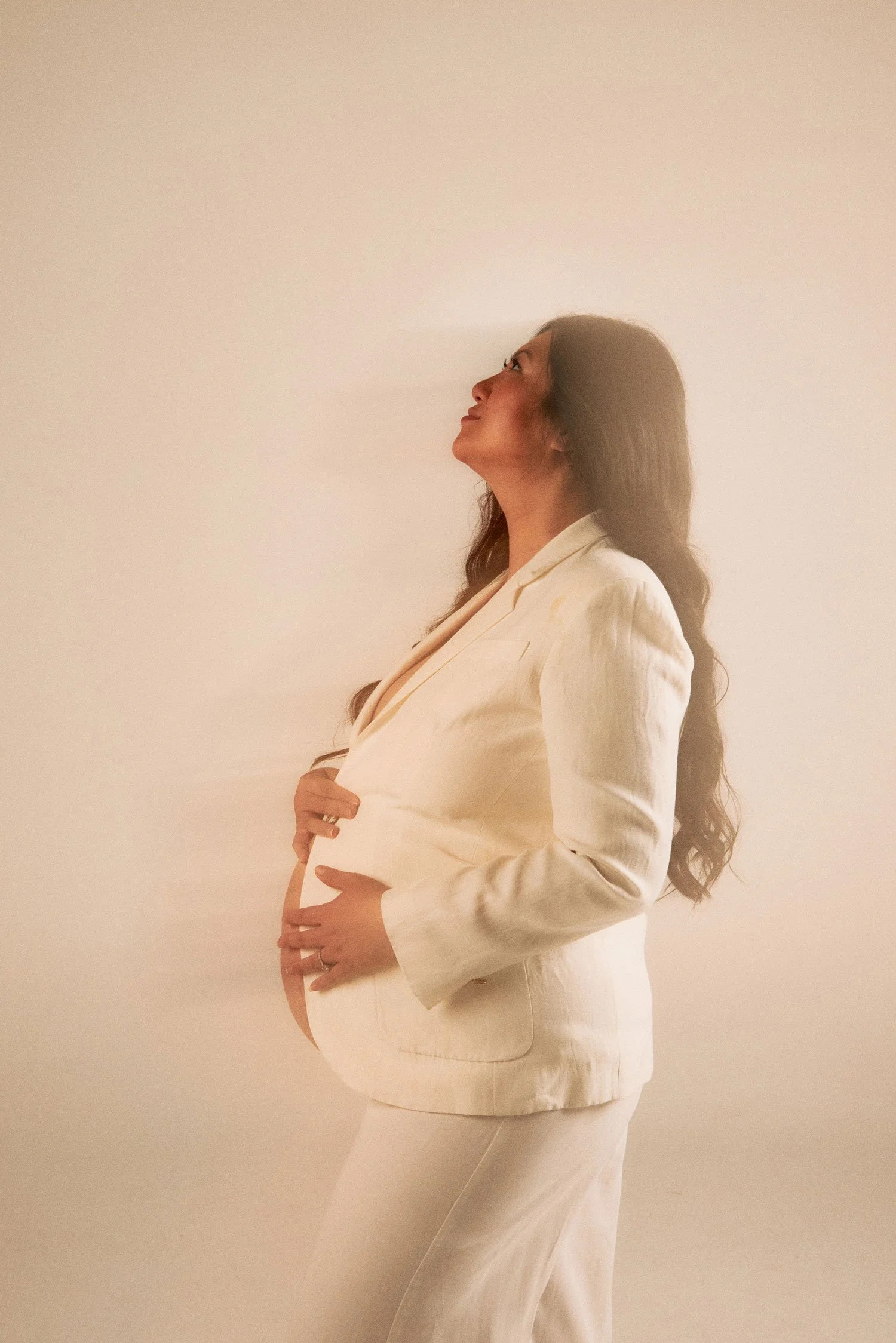 A pregnant woman in a white suit posing against a plain light-colored background, with her hands gently resting on her belly and looking upwards.