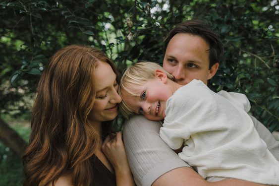 Family of three outdoors, with a woman and man holding a young girl, smiling and enjoying time together among green trees.