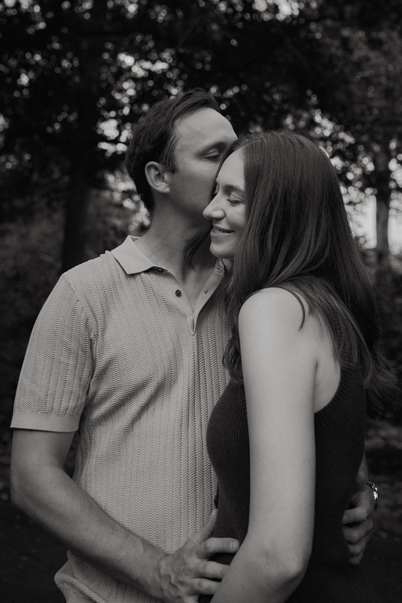 A black and white photo of a man kissing a woman on the forehead while holding her waist, both smiling and standing outdoors with trees in the background.
