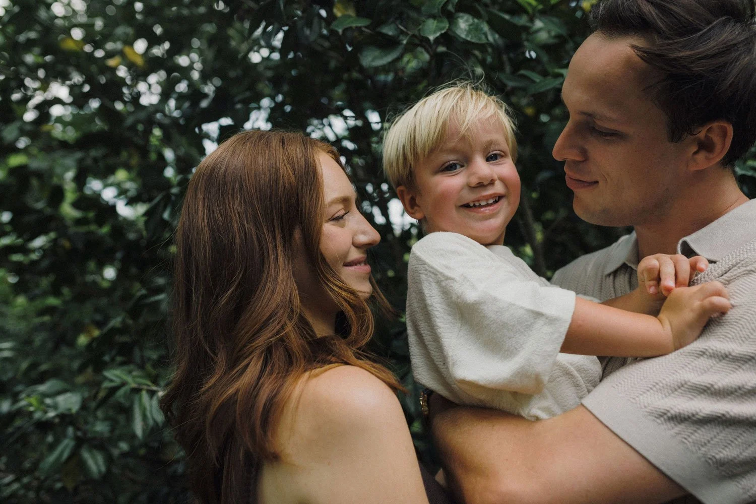 A family of three, a woman, a man, and a young boy, smiling and enjoying a moment together in a lush outdoor setting.