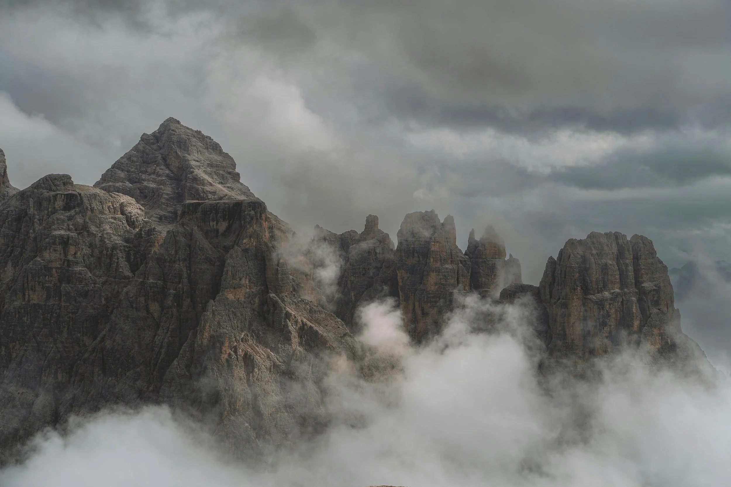 A rugged mountain range with jagged peaks emerging through clouds and mist under a cloudy sky.