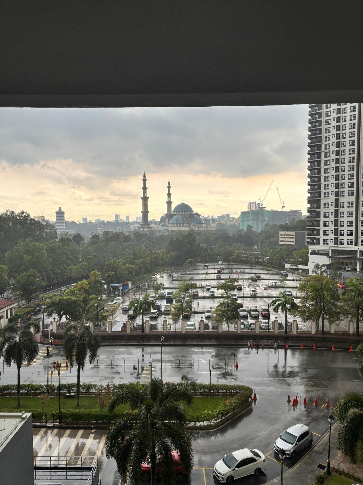 Cityscape view with rain-soaked streets, palm trees, cars, a parking lot, a mosque with minarets, tall residential buildings, and a cloudy sky.