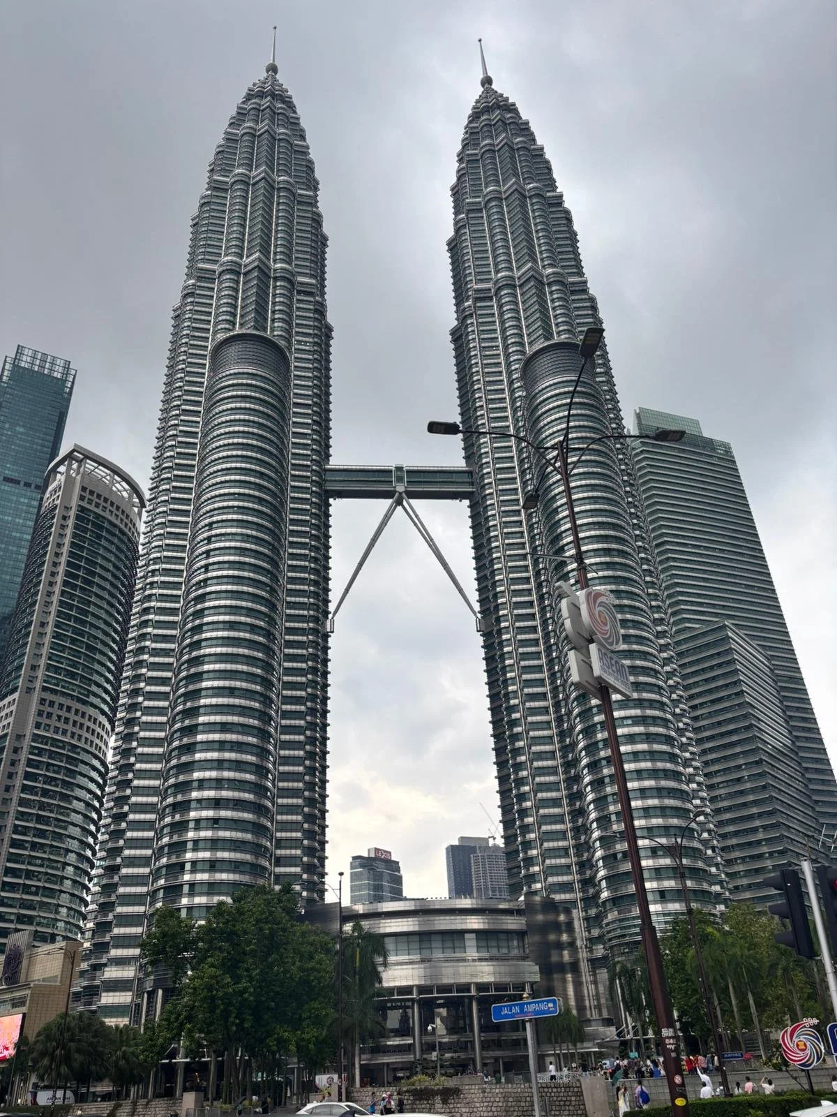 Tall skyscrapers of the Petronas Towers in Kuala Lumpur, Malaysia, connected by a sky bridge, with surrounding modern buildings, trees, and a street scene in foreground.