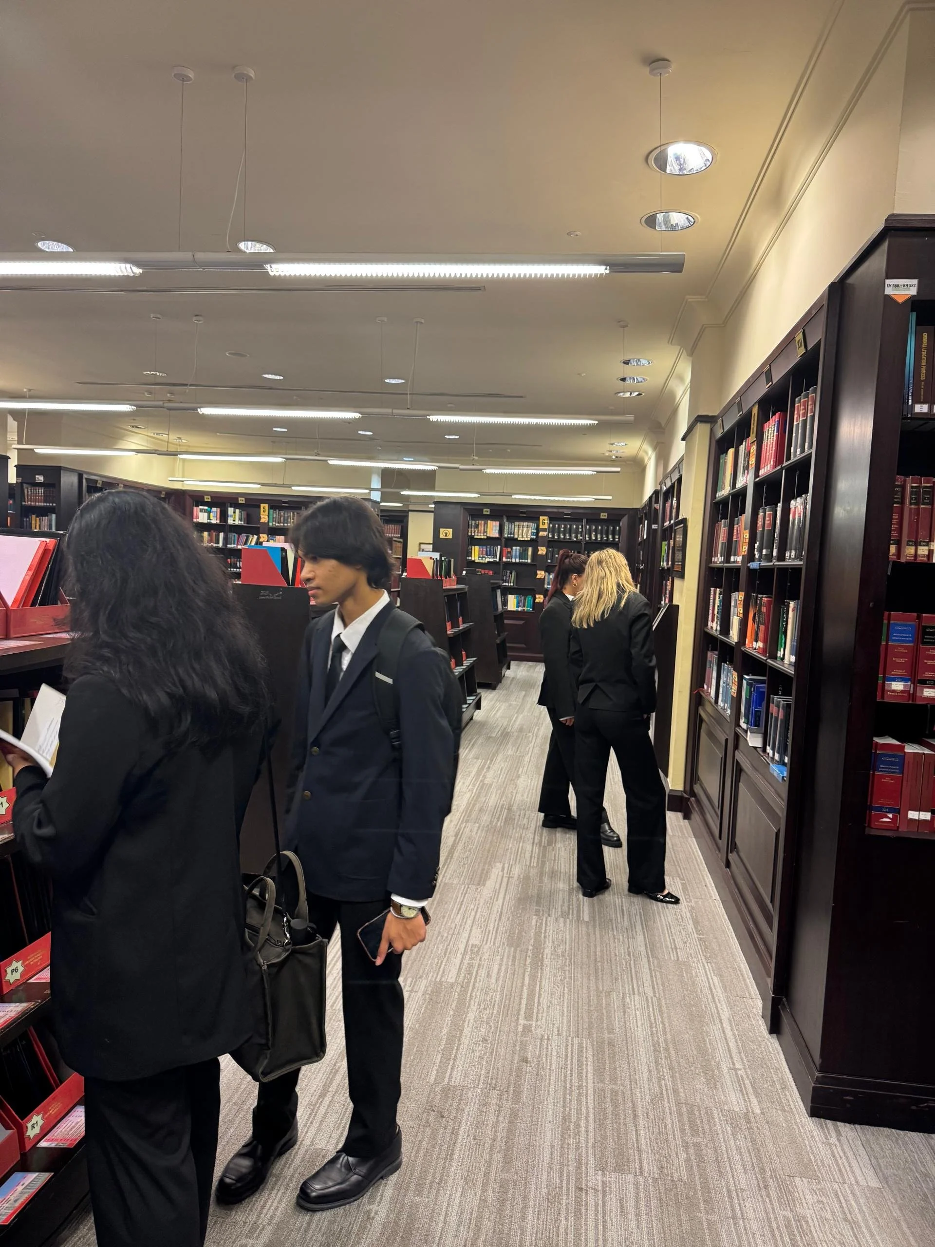 People browsing books in a library with dark wooden bookshelves and bright ceiling lights.