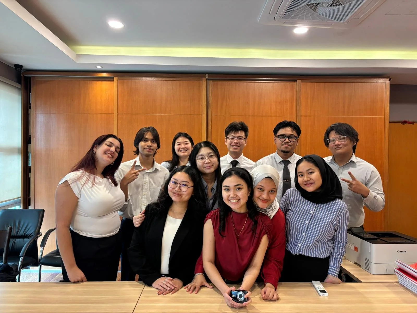 Group of ten diverse young professionals in business attire posing together in a conference room with wooden paneled wall background.