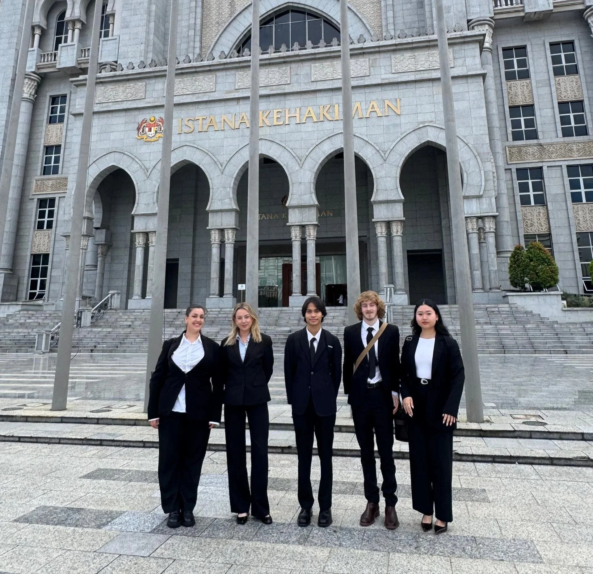 Five young adults in formal business attire standing in front of a large government building with the words 'Istana Kehakiman' on it.