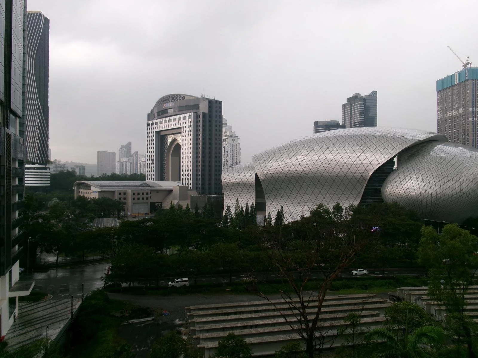 Cityscape with modern buildings, including a distinctive curved silver architectural structure and a tall building with a large arch and patterned facade, under an overcast sky.