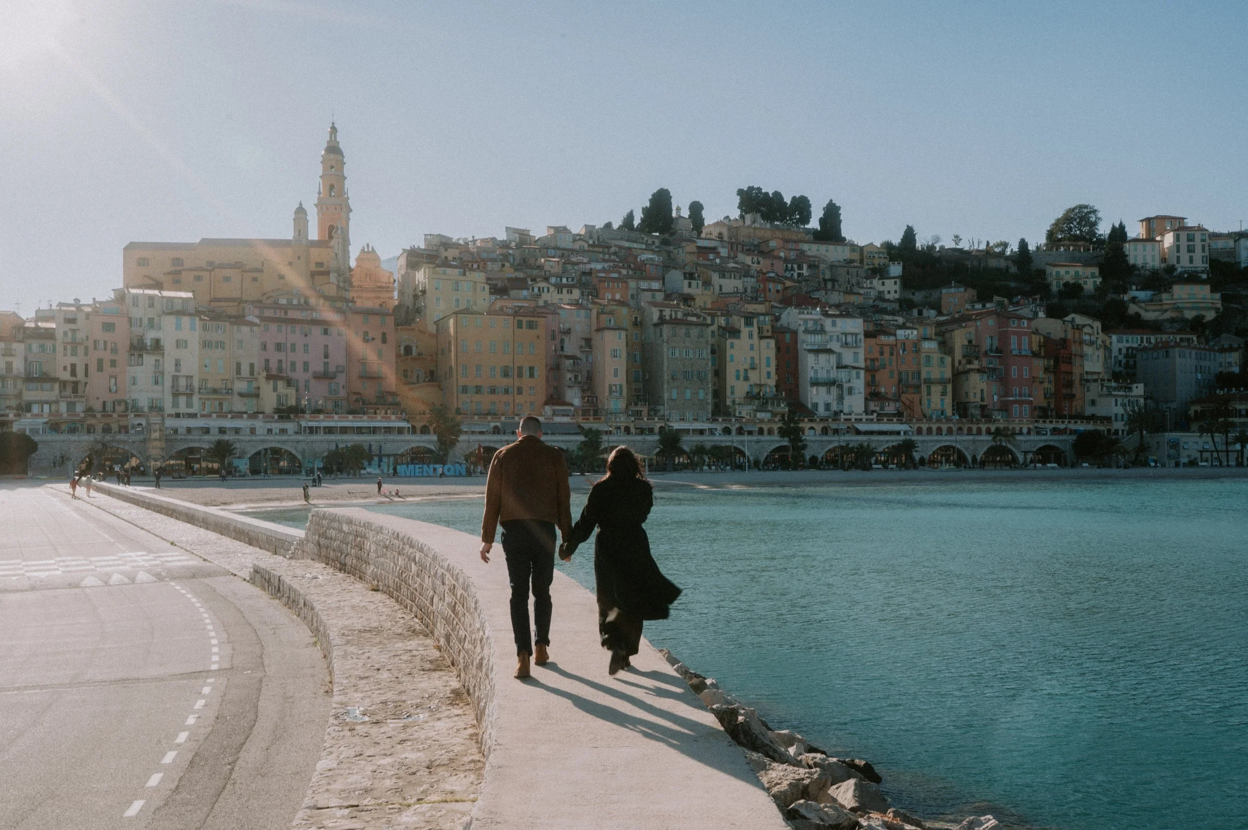 A man and woman walk hand-in-hand along a paved waterfront promenade with a stone barrier on the La Mortella hillside with colorful buildings and a church tower in the background.