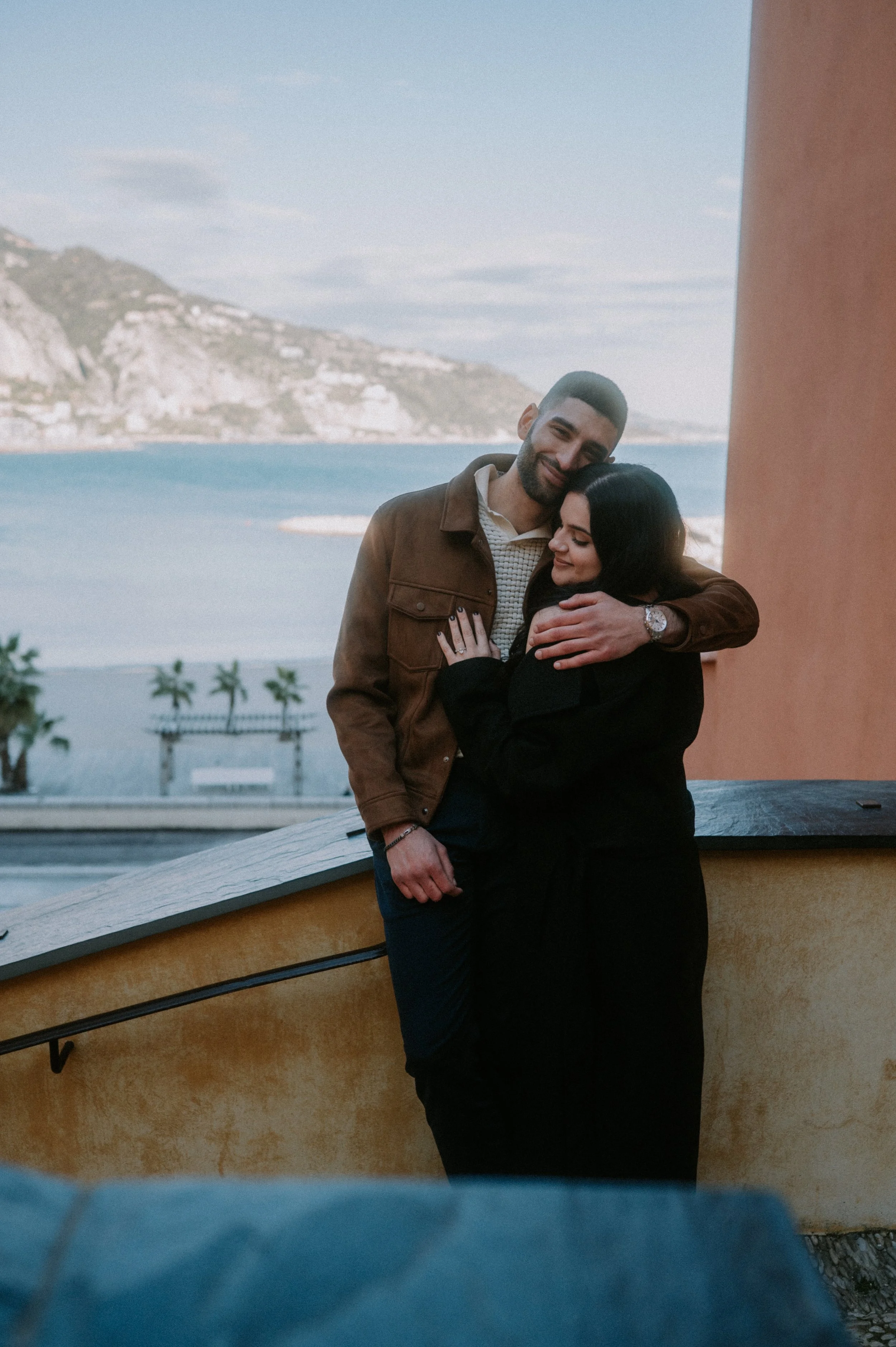A man and woman hugging each other outdoors by a yellow wall with a scenic background of the ocean, mountains, and cloudy sky.