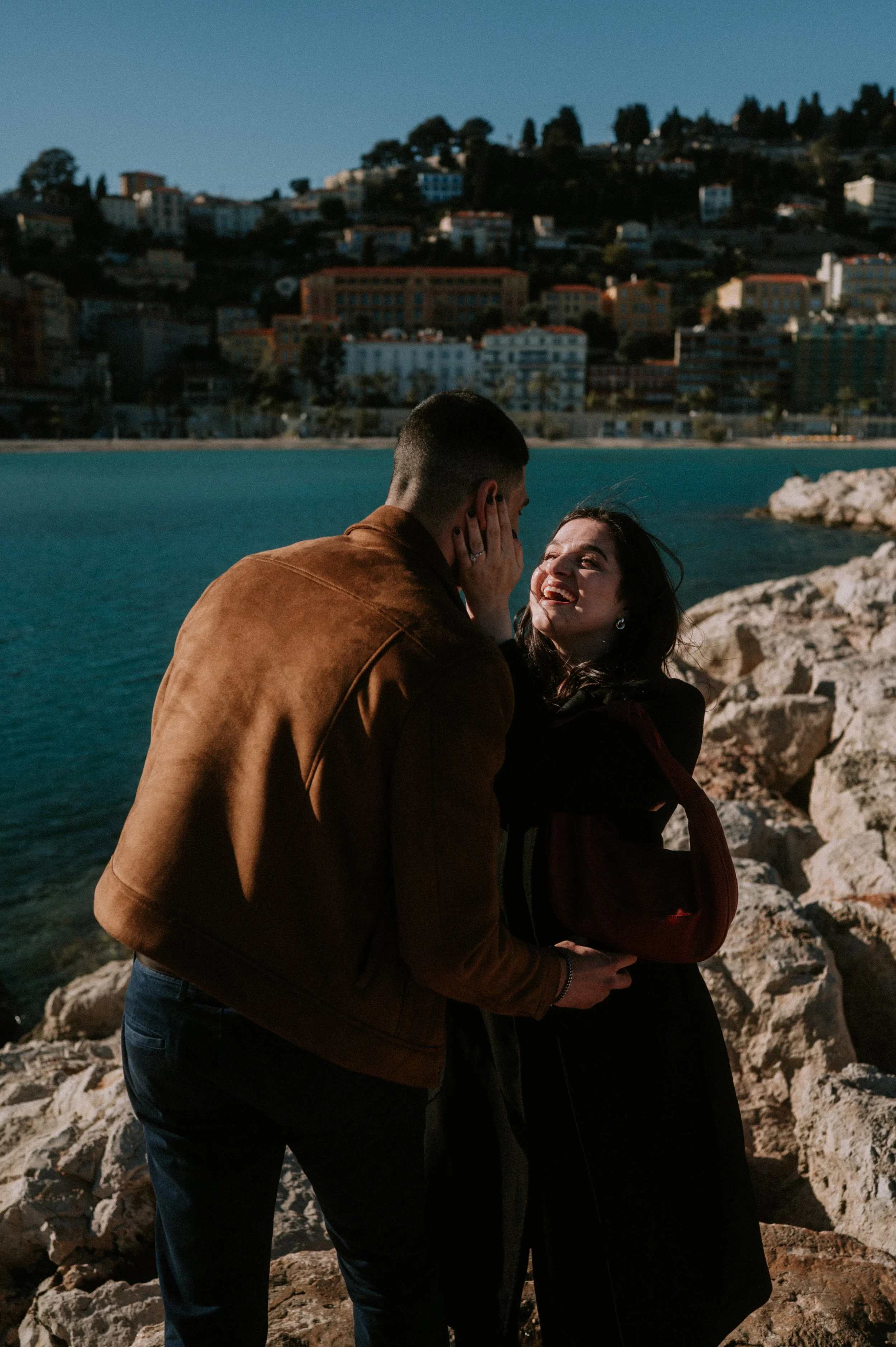 A couple stands on a rocky shoreline with a cityscape on a hillside in the background, embracing and smiling at each other during sunset.