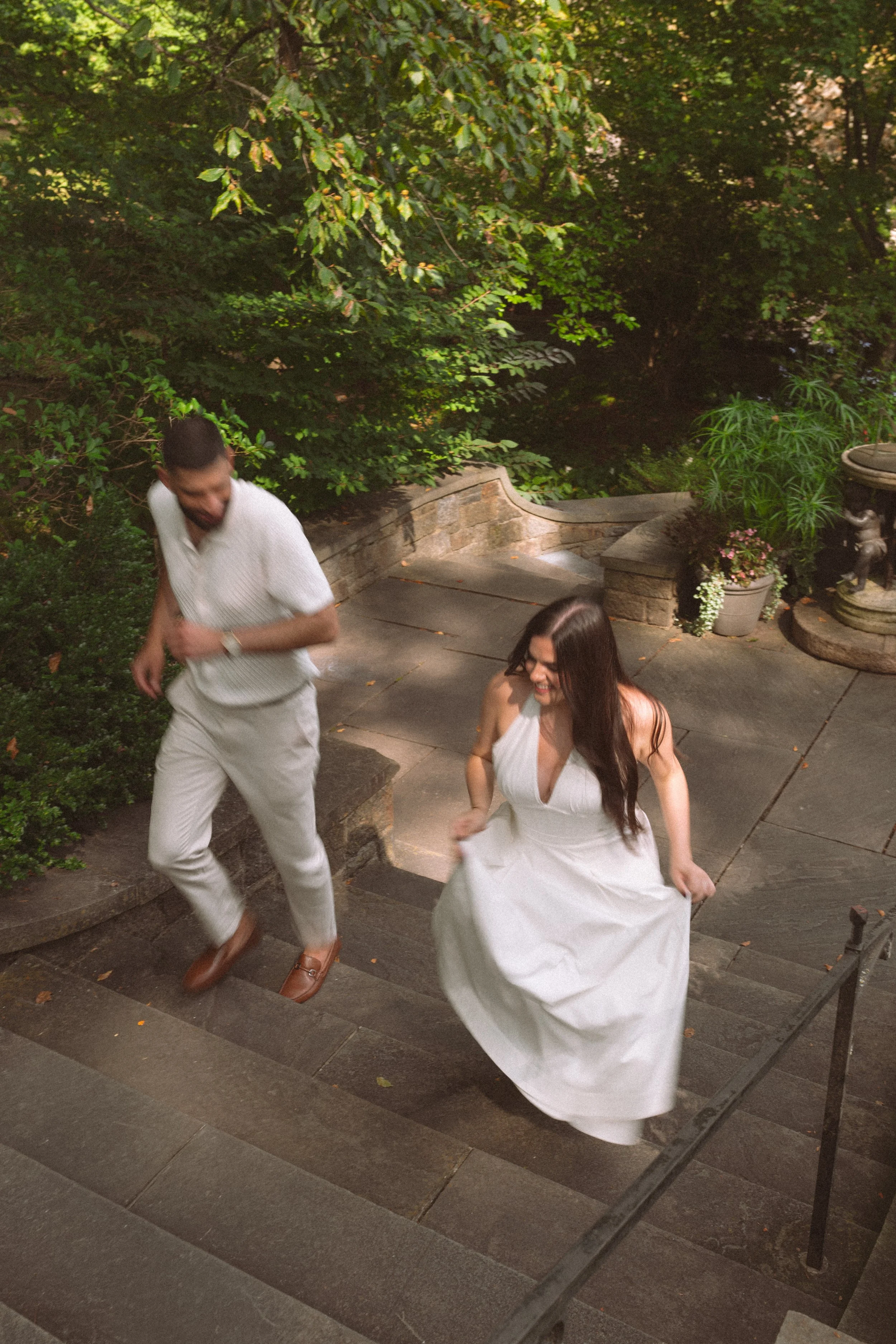 A man and a woman are descending outdoor stairs surrounded by greenery, with the woman holding up her dress to avoid tripping.