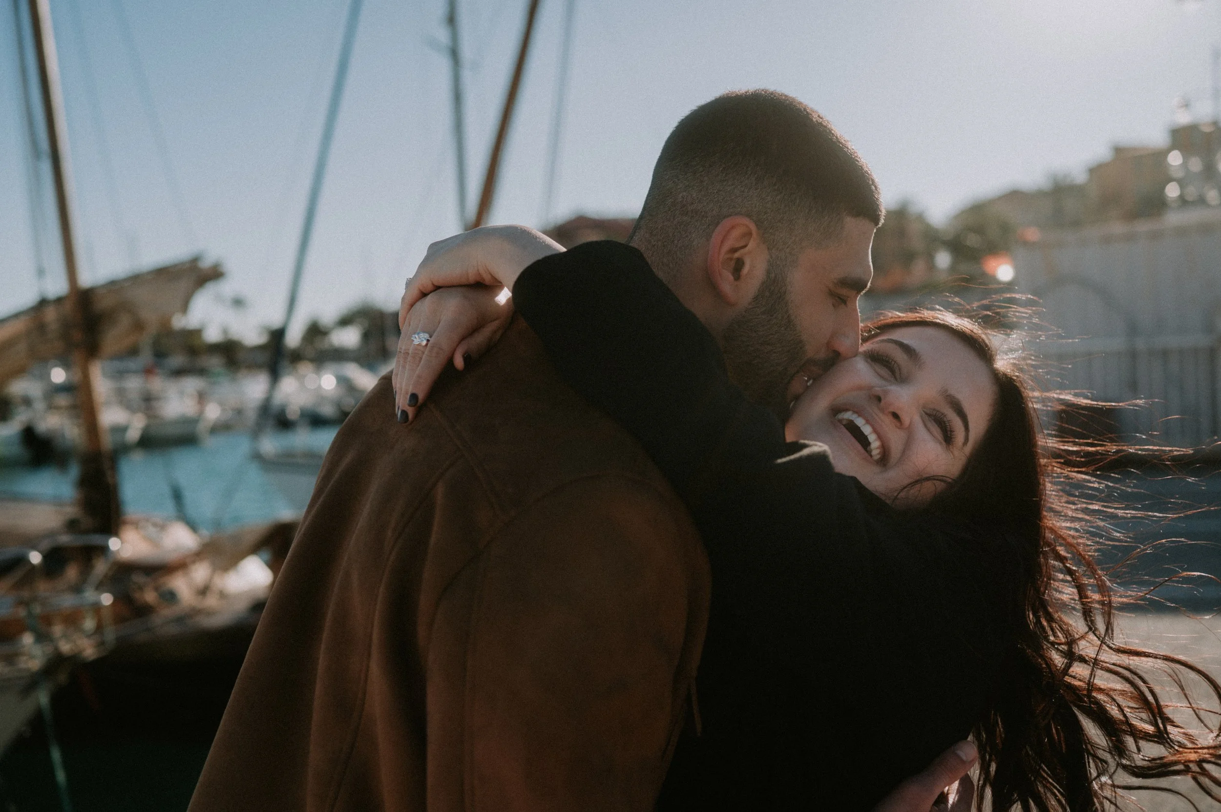 A happy couple hugging outdoors near a marina with boats, during late afternoon sunlight.