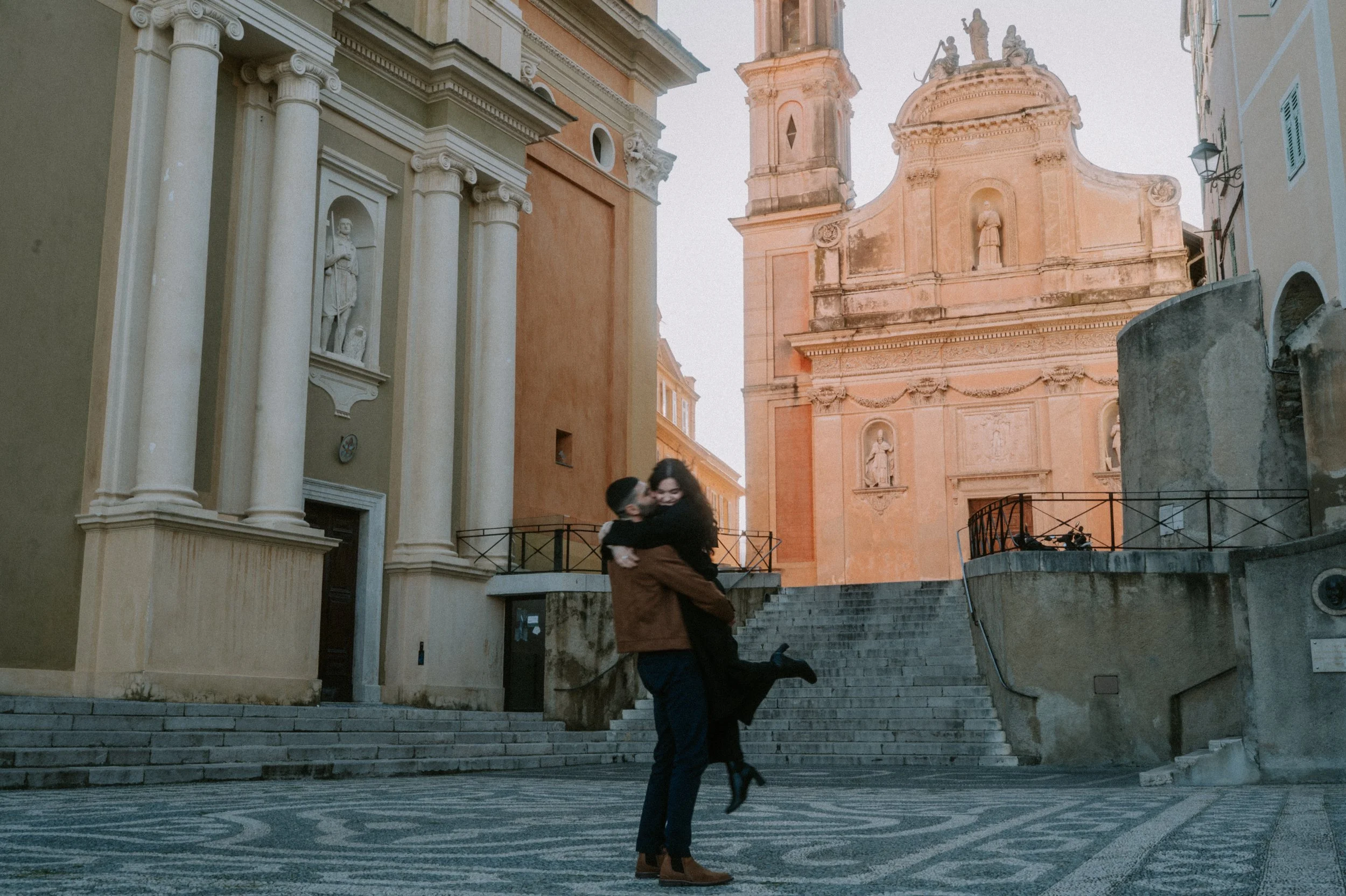 A couple dancing in a European town square surrounded by historic buildings with ornate facades and statues, during late afternoon or early evening.