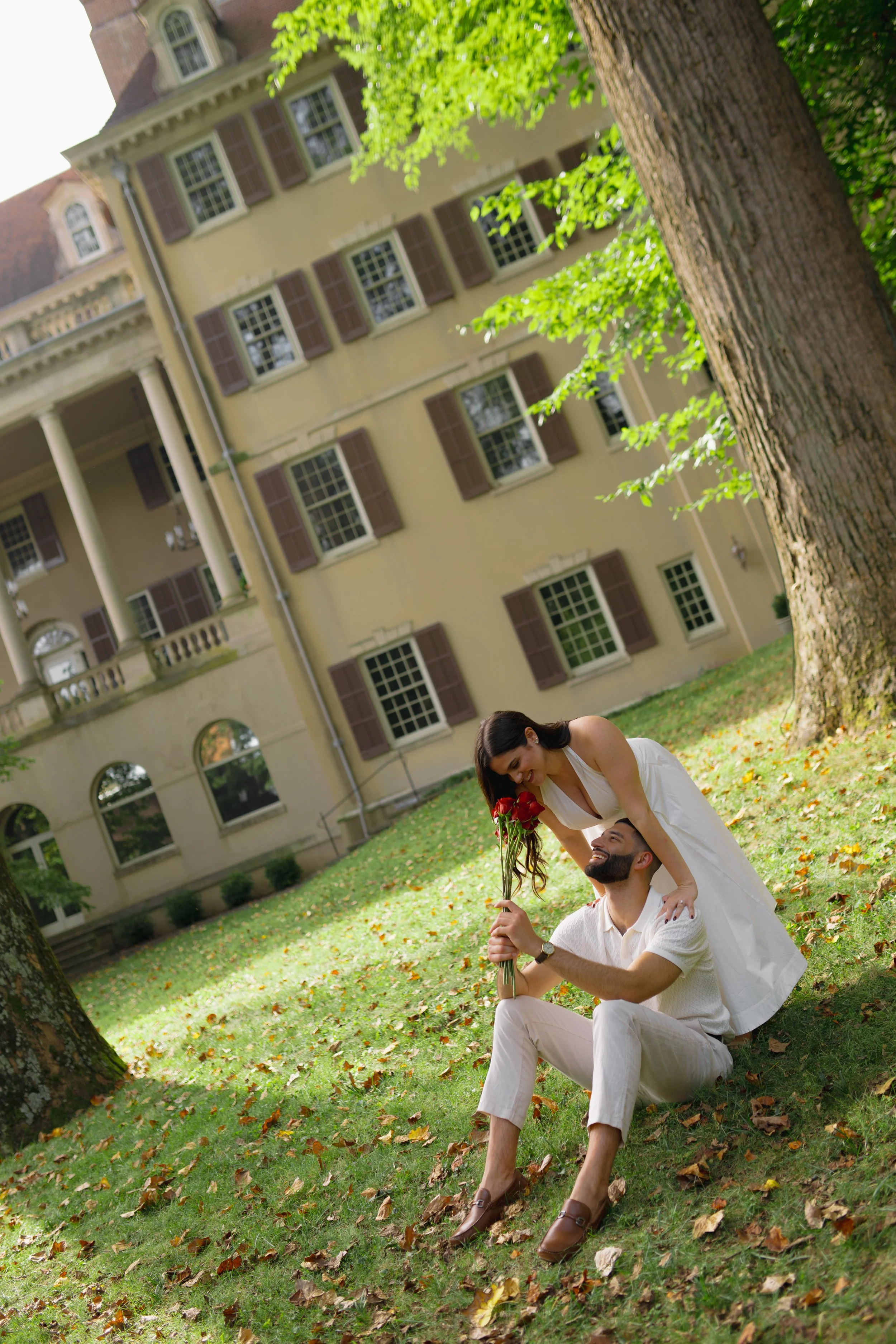 A woman in a white dress sitting on the grass and a man in white clothes kneeling beside her, holding a bouquet of red roses in front of a large tree with green leaves. A large yellow house with multiple windows and shutters is in the background.