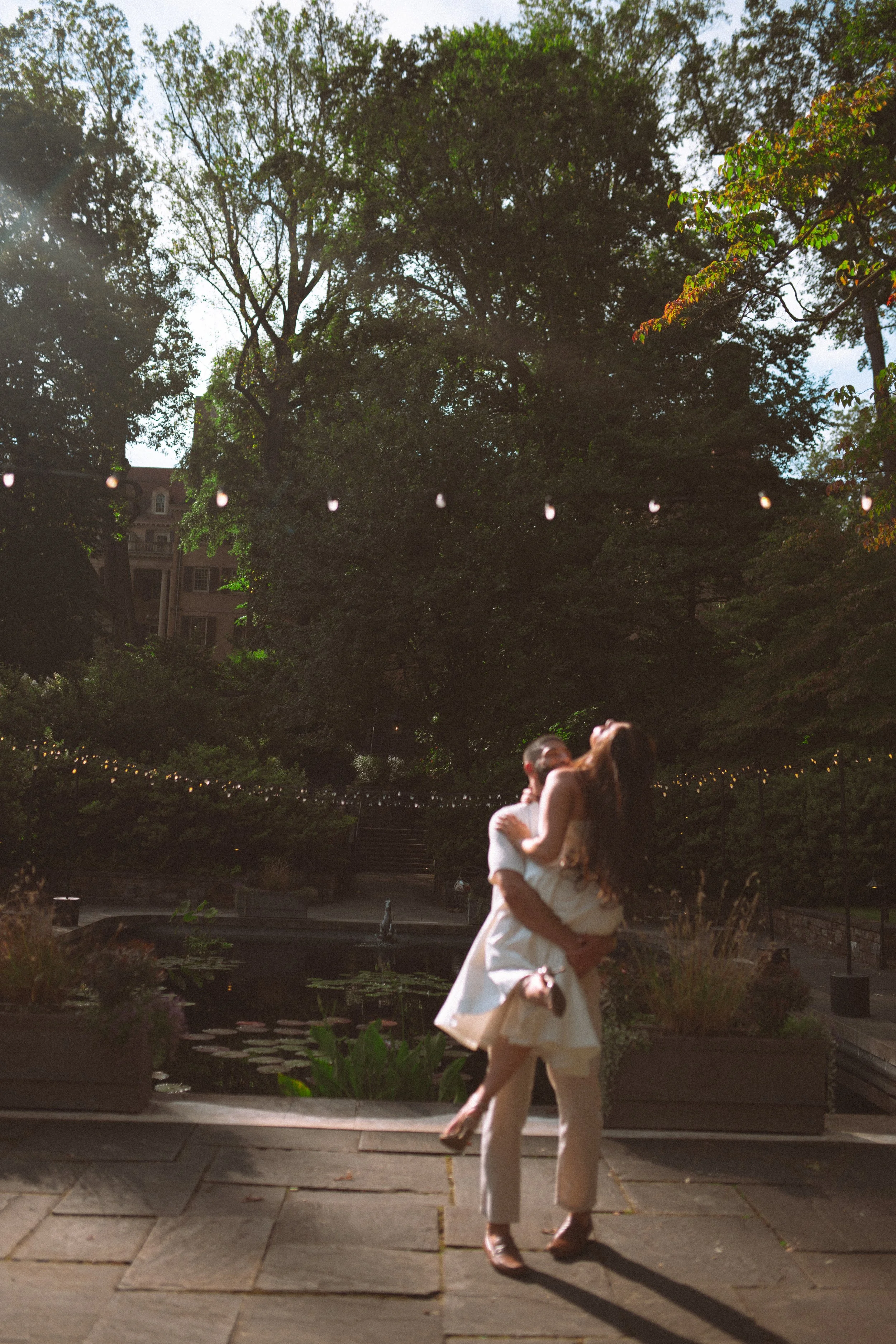 A man and woman are dancing closely outdoors during dusk or evening, with trees, string lights, and a building in the background.
