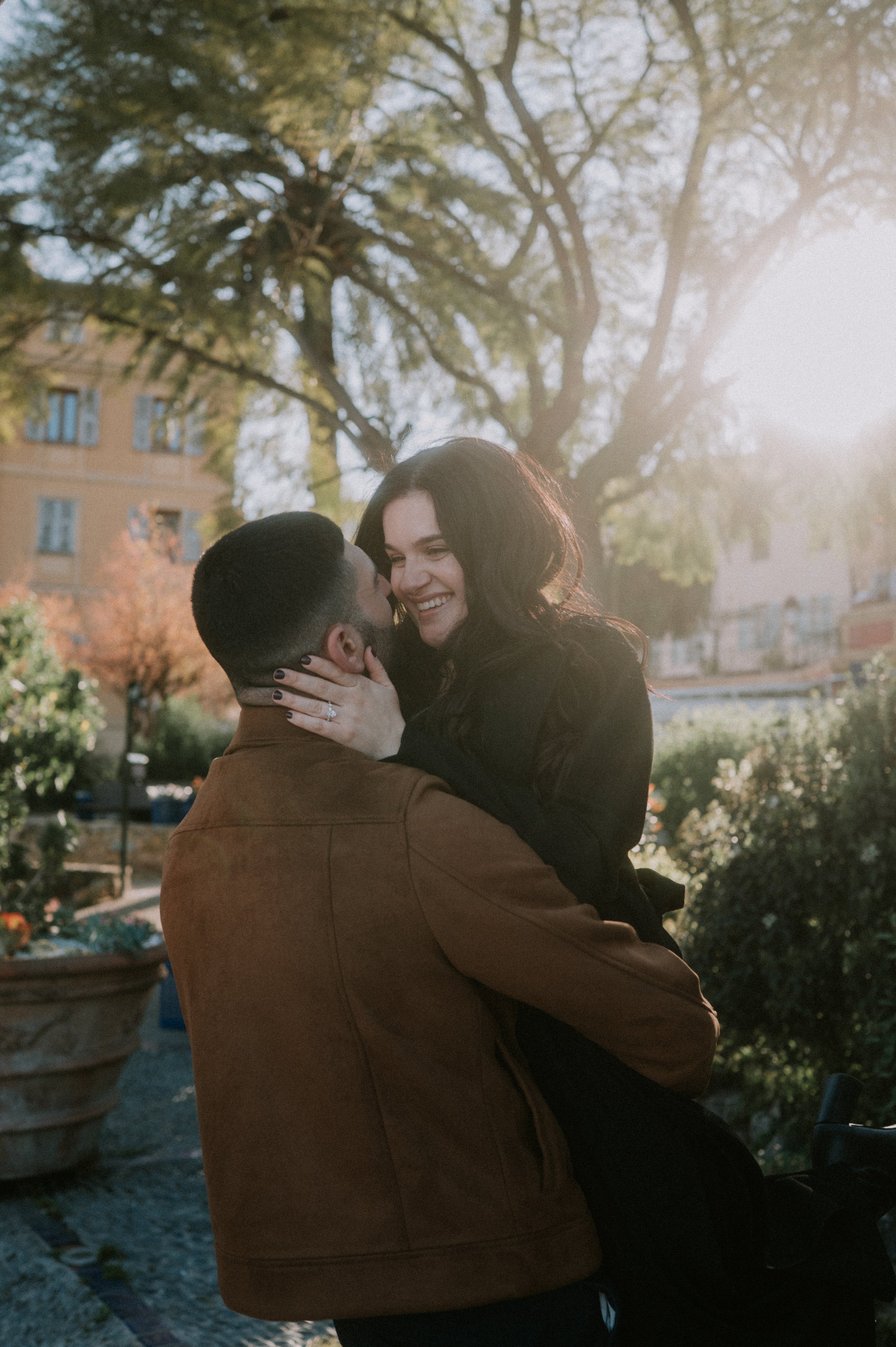 A couple sharing an embrace outdoors in a park during sunset, with trees and buildings in the background.