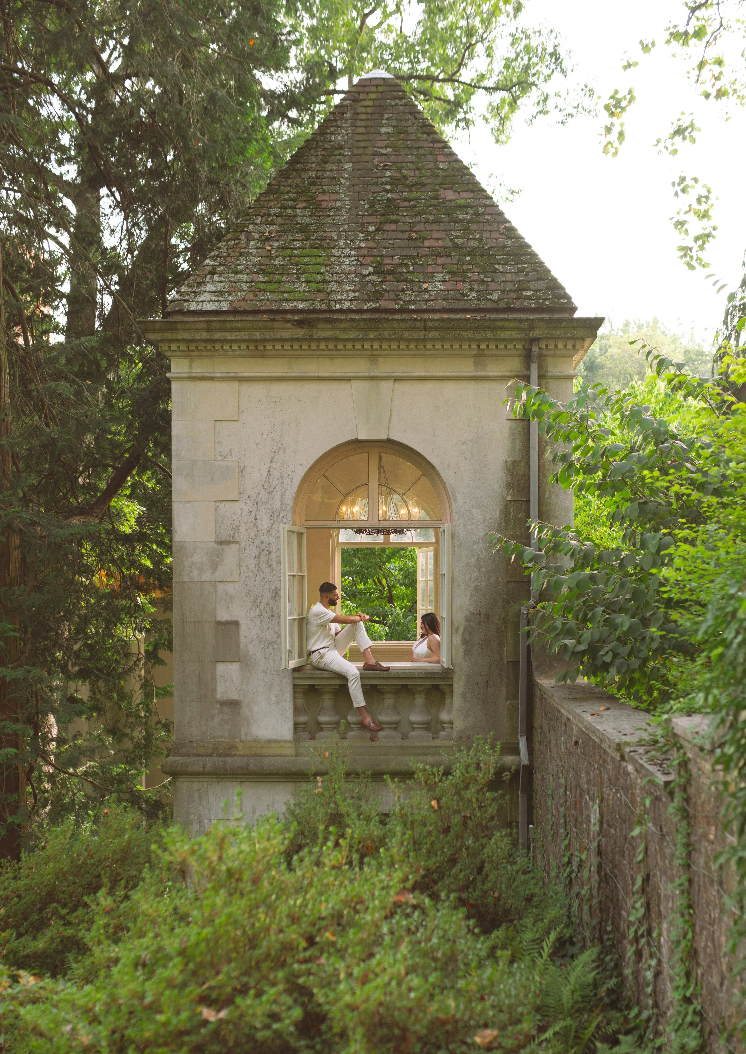 A man and woman sitting in an open window of a stone building surrounded by greenery, with trees and bushes visible around.