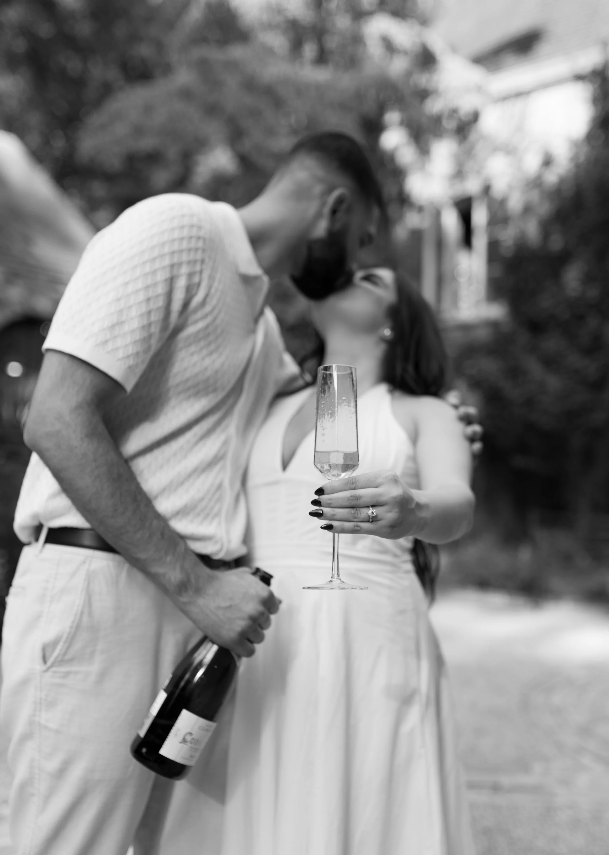 A couple kissing outdoors, with the woman holding a glass of champagne and a wine bottle in her hand, both dressed in white. Black and white photo.