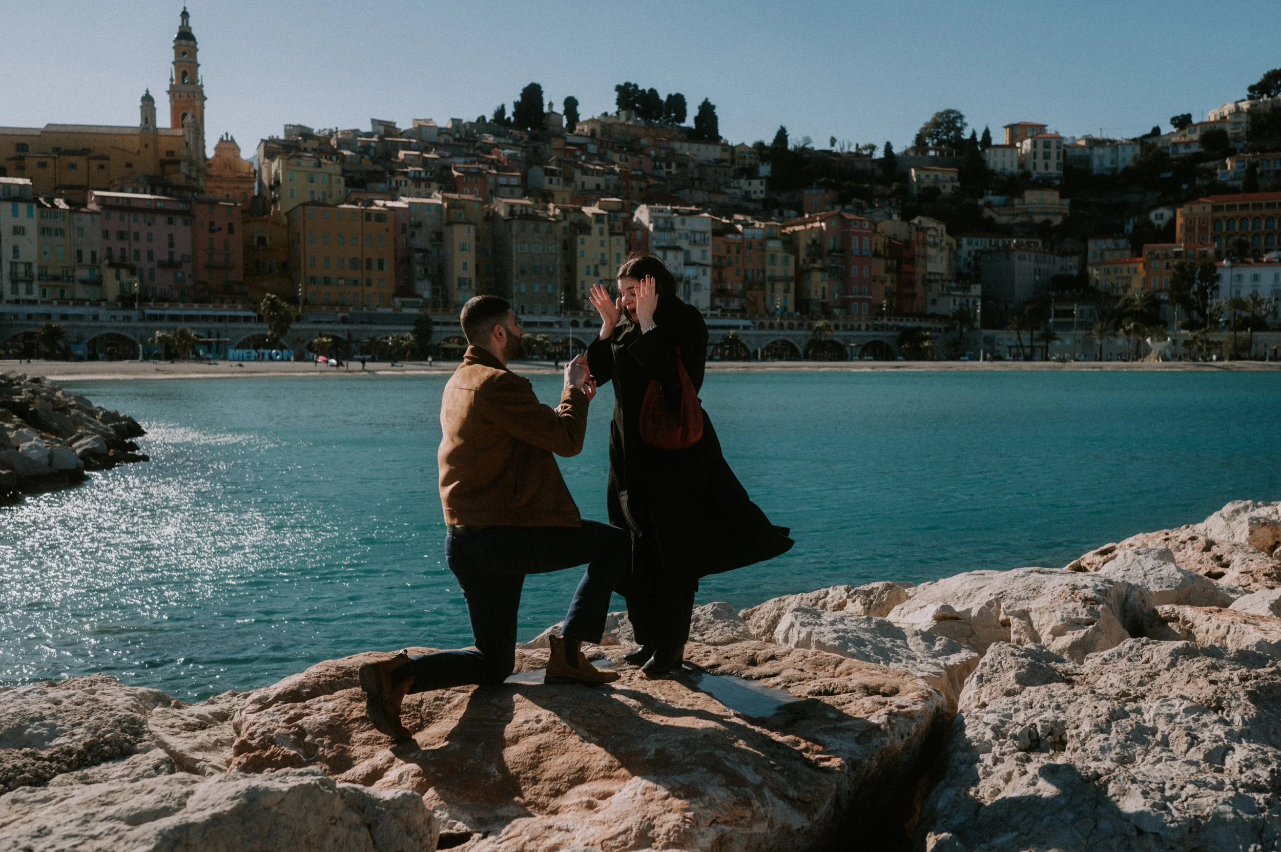 A man on one knee proposing to a woman along a rocky shoreline with a cityscape of hillside colorful buildings in the background.