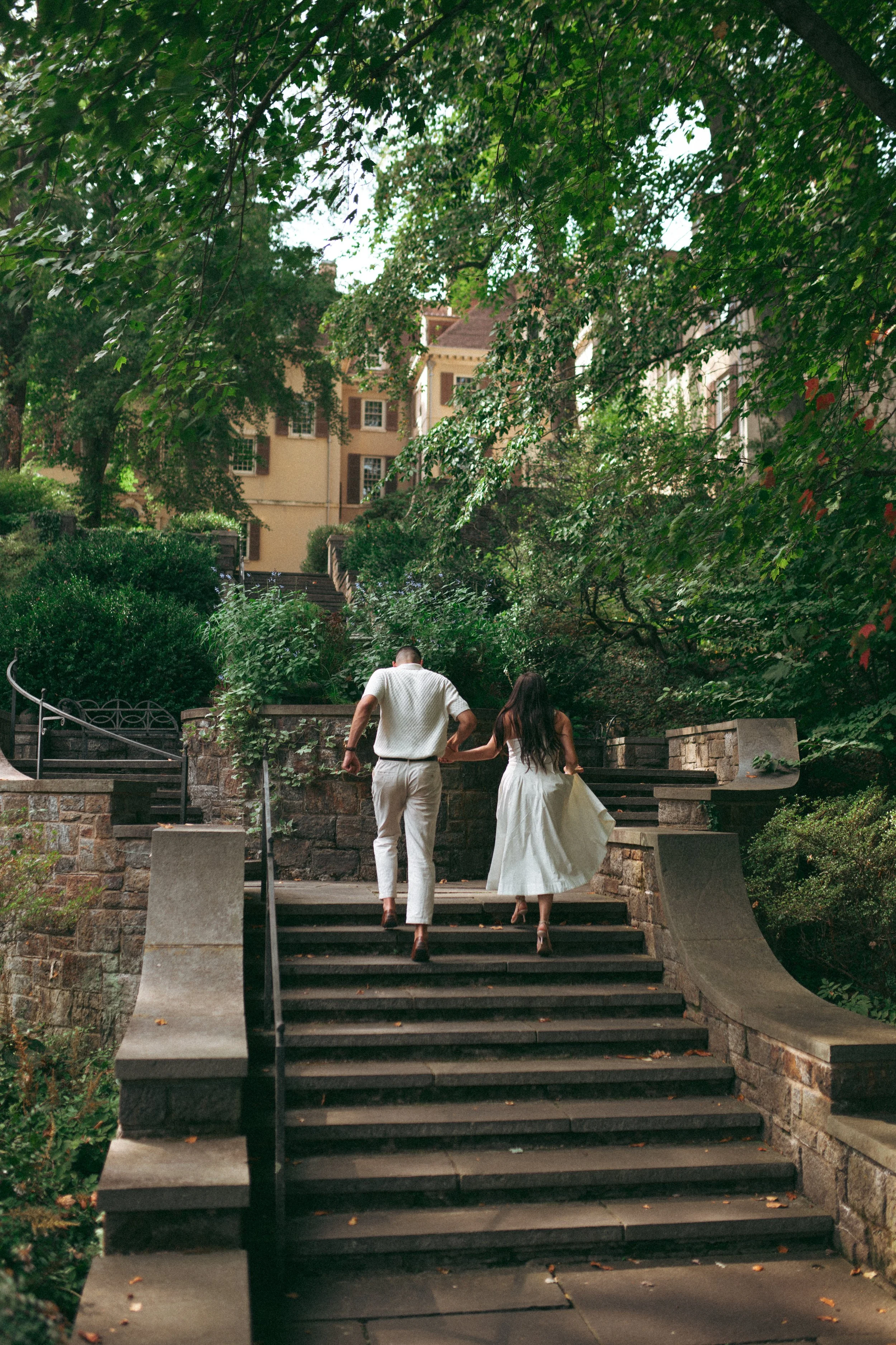 A couple walking up city stairs in a green park area with trees and shrubs, in front of a tan-colored building with multiple windows.