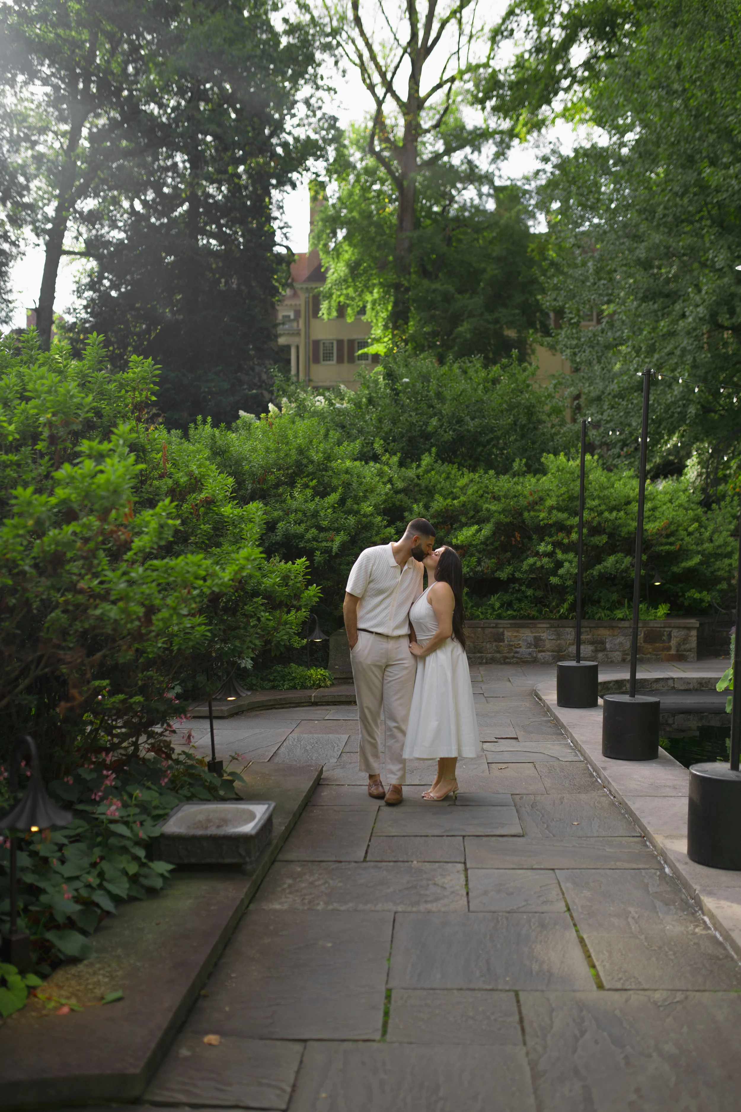 A couple sharing a kiss in a lush outdoor garden, surrounded by greenery and trees, with a building in the background and string lights hanging nearby.