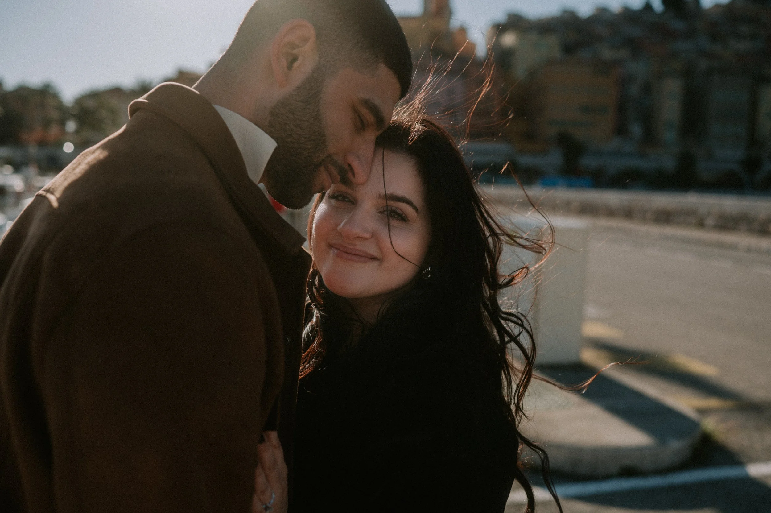 A young man with a beard and short hair leaning his forehead against a young woman with long dark hair, smiling softly. They are outside with a blurred background of trees and a parked boat. The sunlight illuminates their faces and hair.