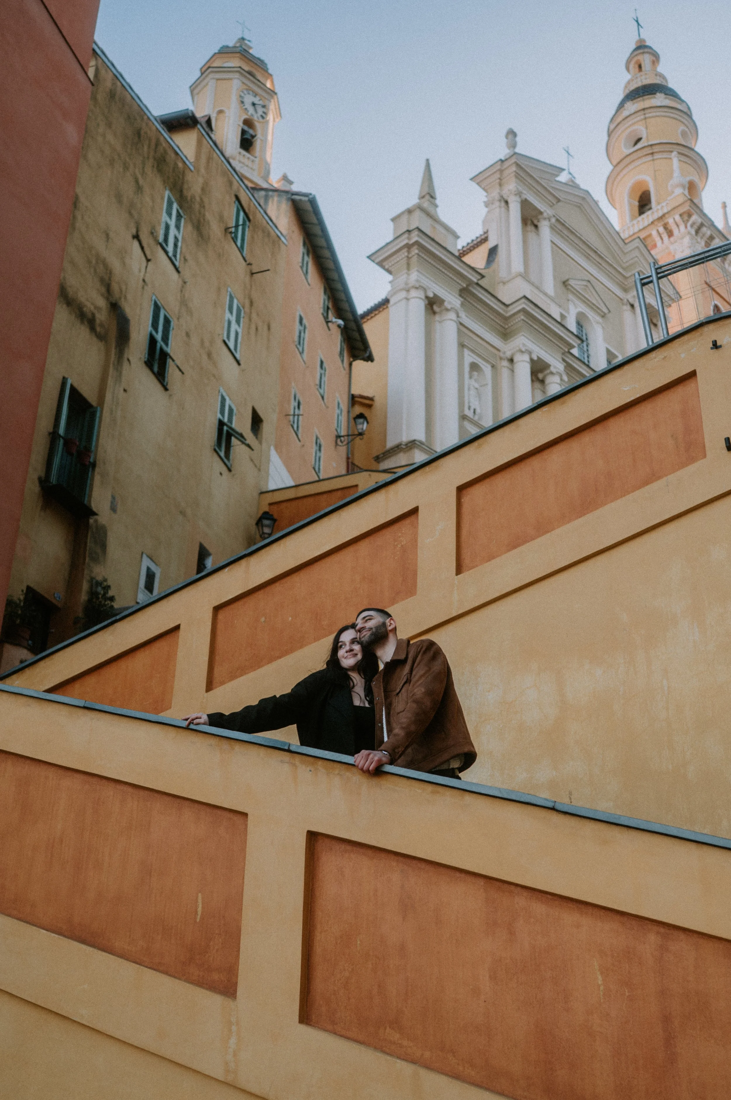 A couple stands on a yellow balcony, with a historic cityscape and church towers behind them. The woman has dark hair and is wearing a black coat, and the man has a beard, glasses, and is wearing a brown jacket. They are smiling and looking into the distance.
