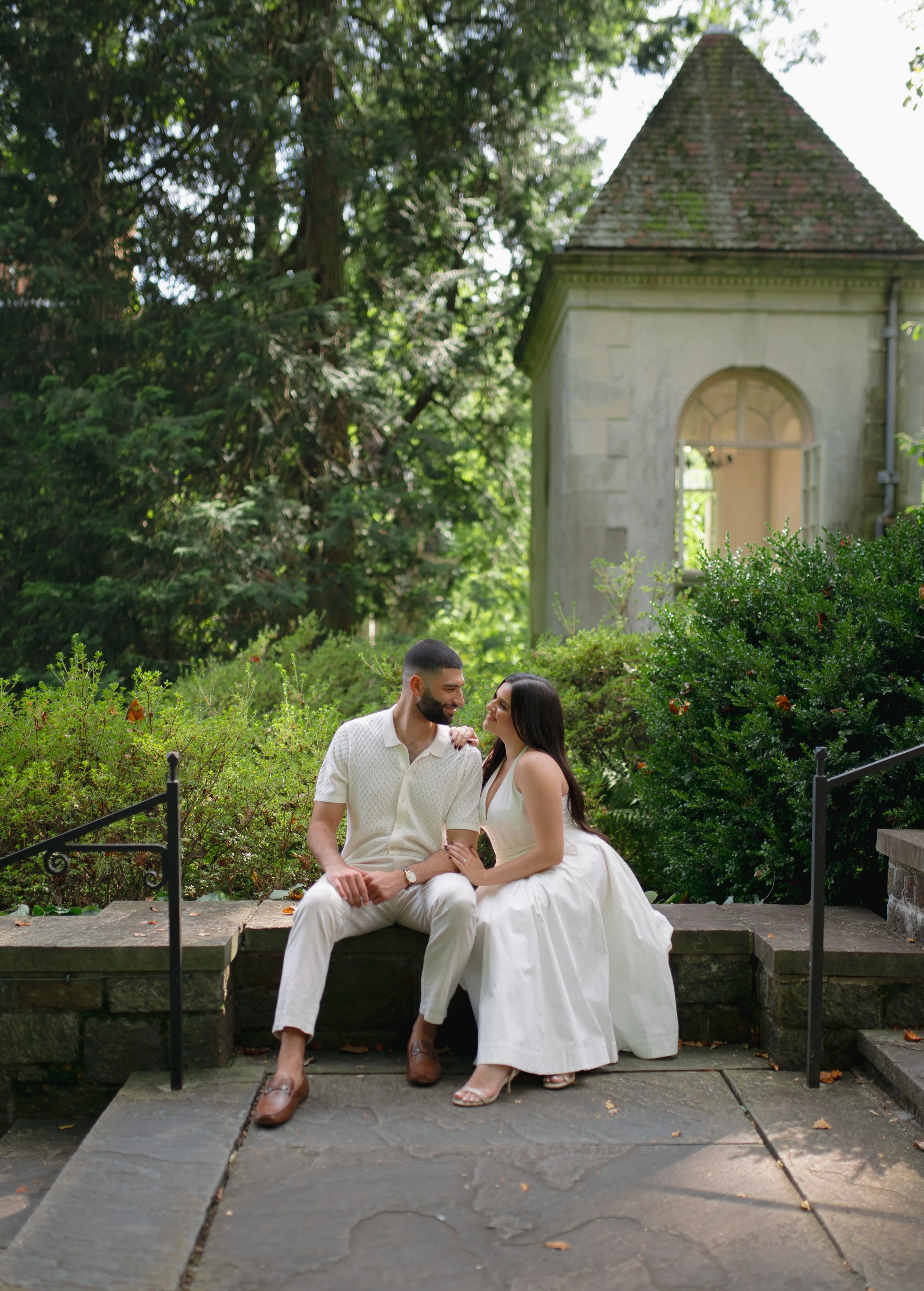A couple sitting on a stone step outdoors, near a lush garden with green bushes and trees, in front of a small stone building with a triangular roof and arched window, smiling and looking at each other.