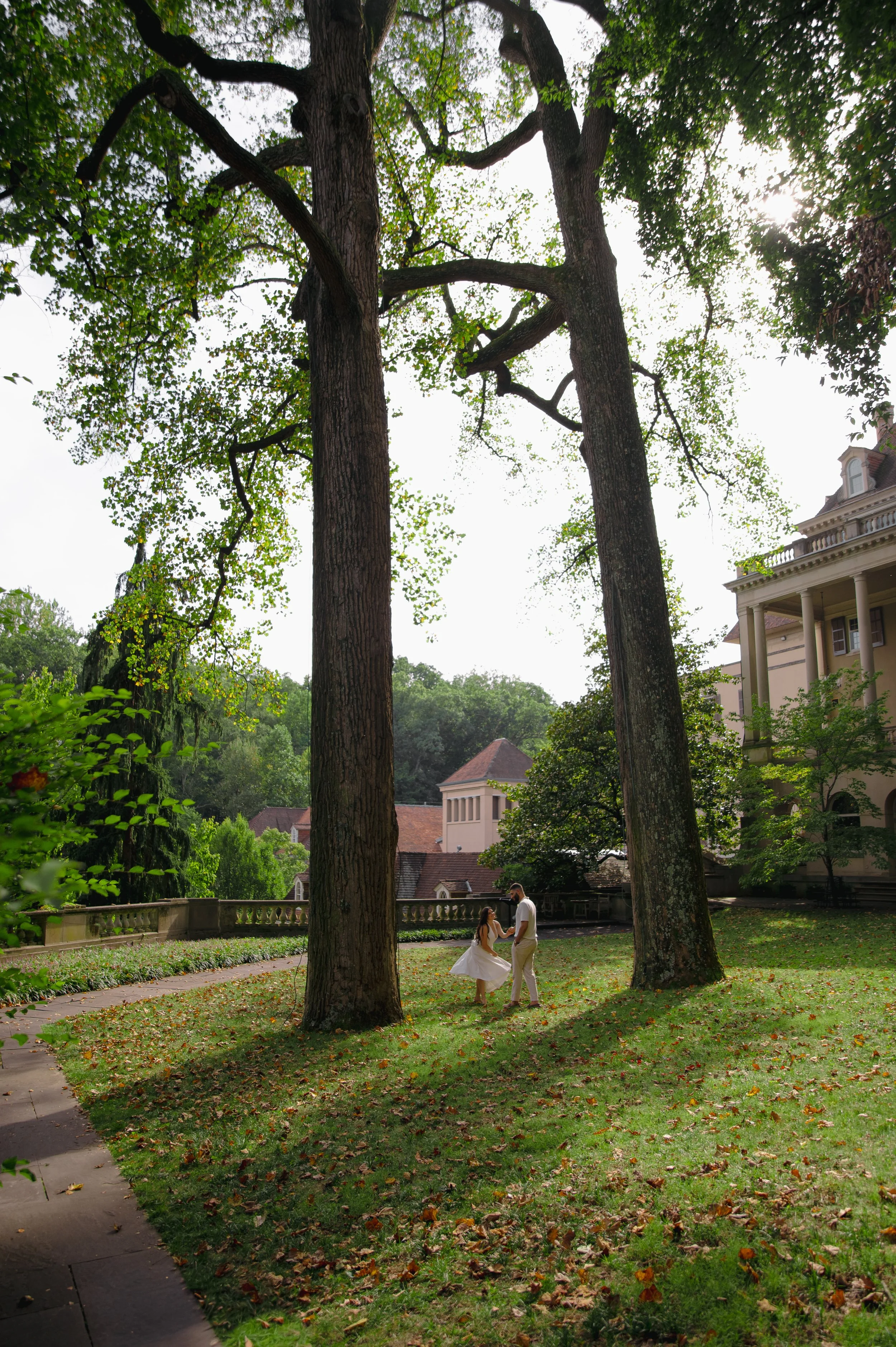 A couple holding hands in a park with tall trees, fallen leaves, and a large house in the background.