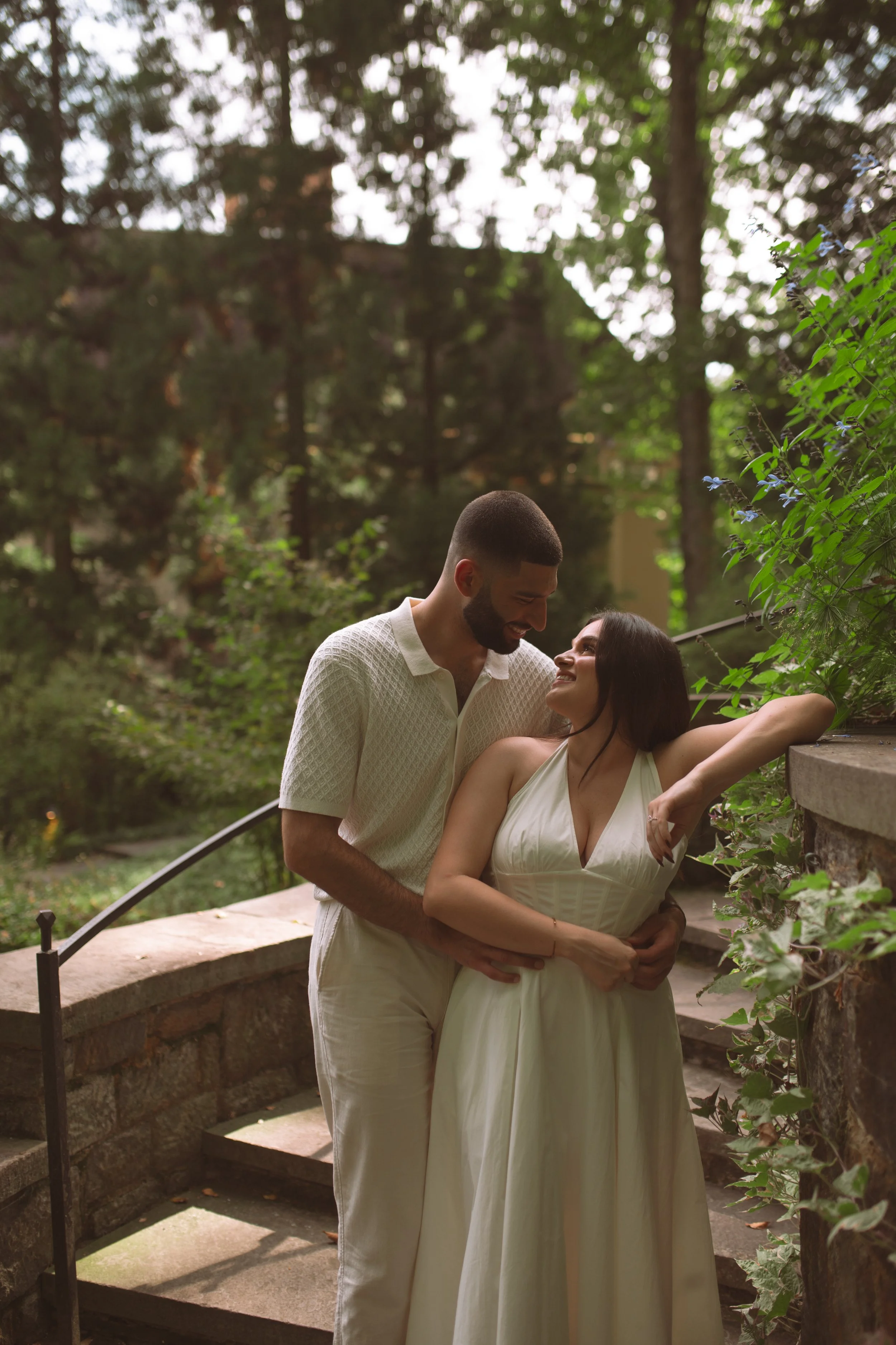 A couple standing close together outdoors in a garden park, smiling and looking at each other, with trees and greenery in the background.