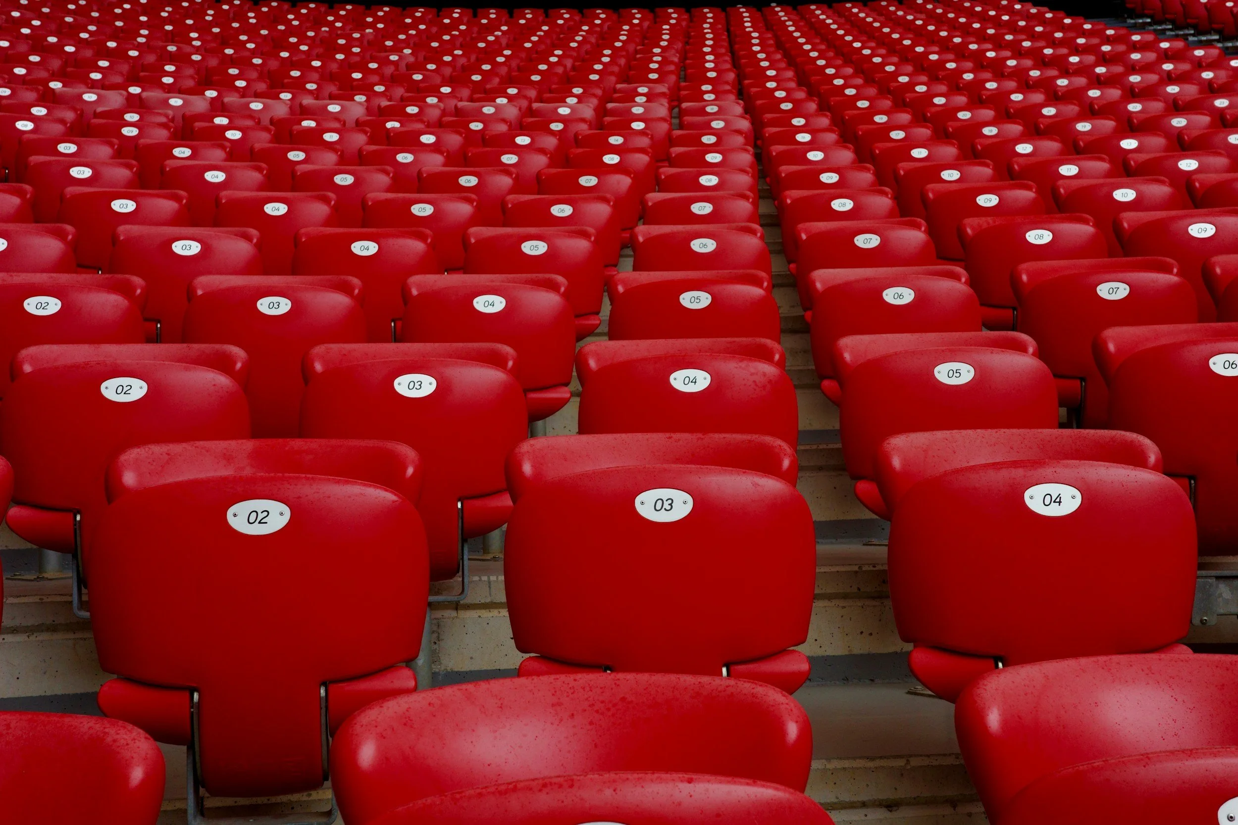 A large stadium or arena filled with rows of red plastic seats with white stickers numbered from 02 to 13.